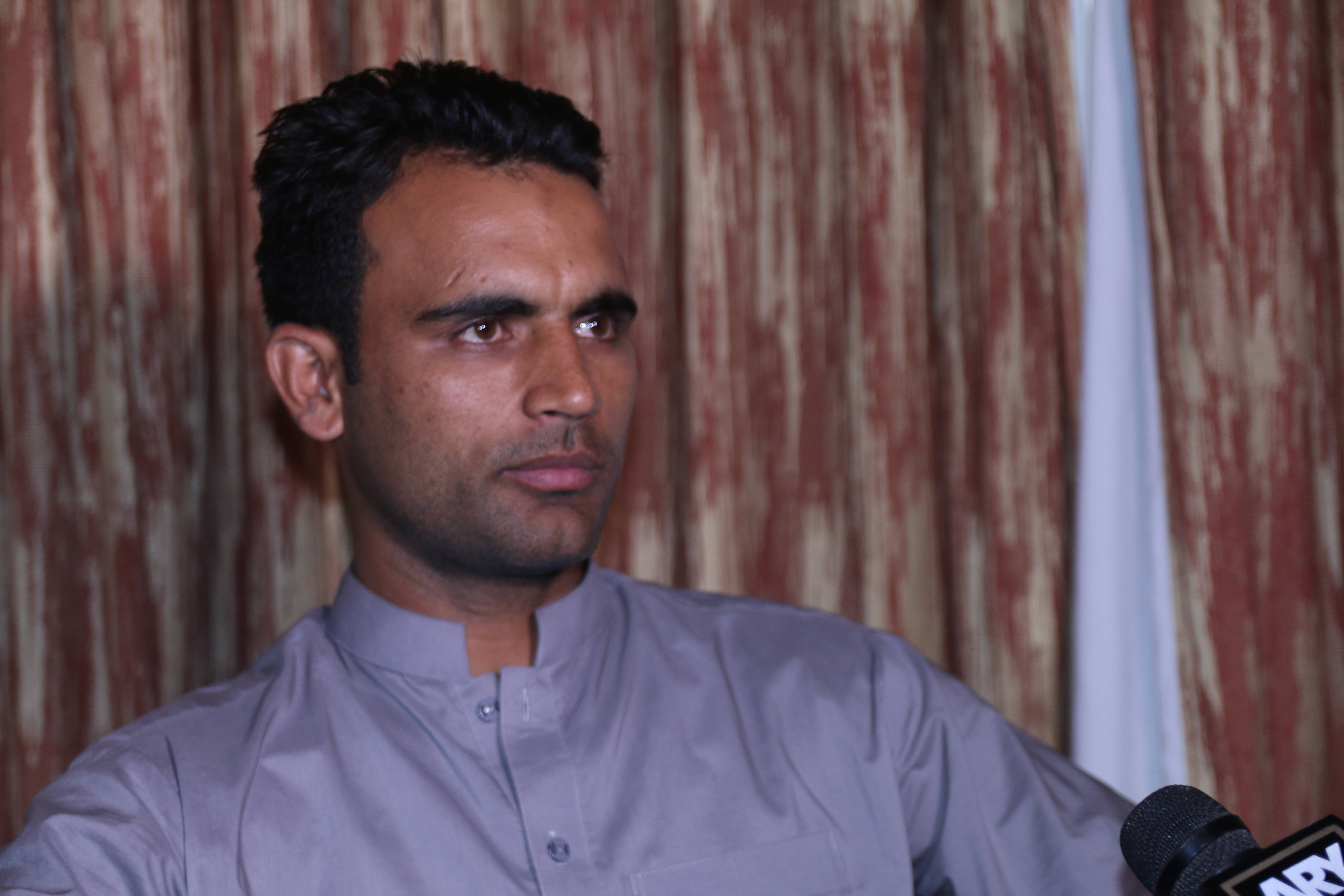 Fakhar Zaman 桌布 壁紙 — 一名身穿灰色傳統襯衫的男子在帶有紋理的紅色窗簾背景前對著麥克風講話。