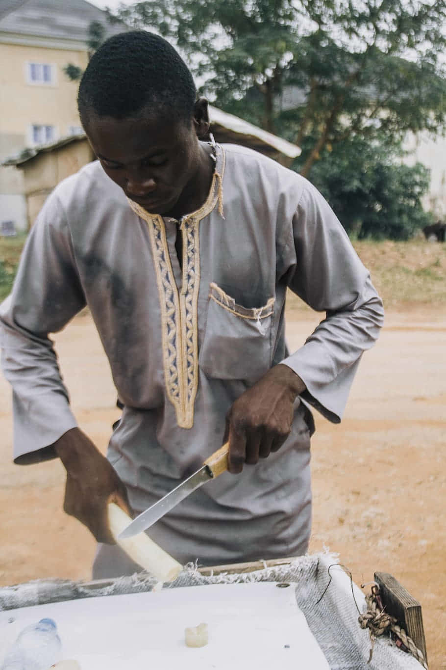Nigeria 桌布 壁紙 — 一名身穿灰色刺繡襯衫的男子，正在戶外的木砧板上小心地切一個白色塊莖。