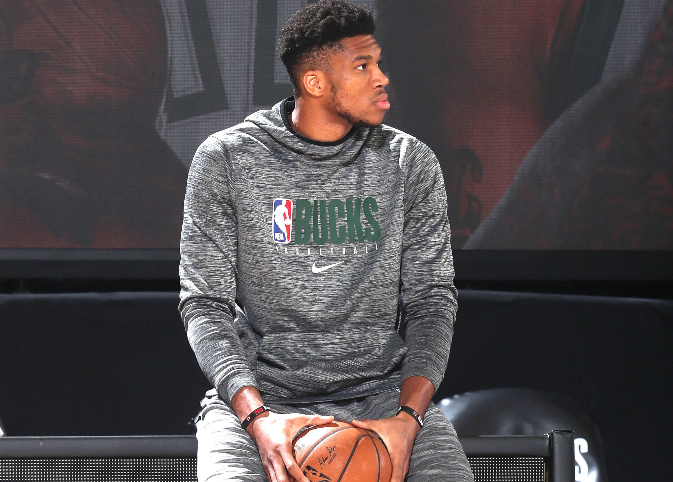 Giannis 桌布 壁紙 — 一名身穿灰色密爾瓦基公鹿連帽衫的籃球運動員坐著，手持籃球，神情專注。