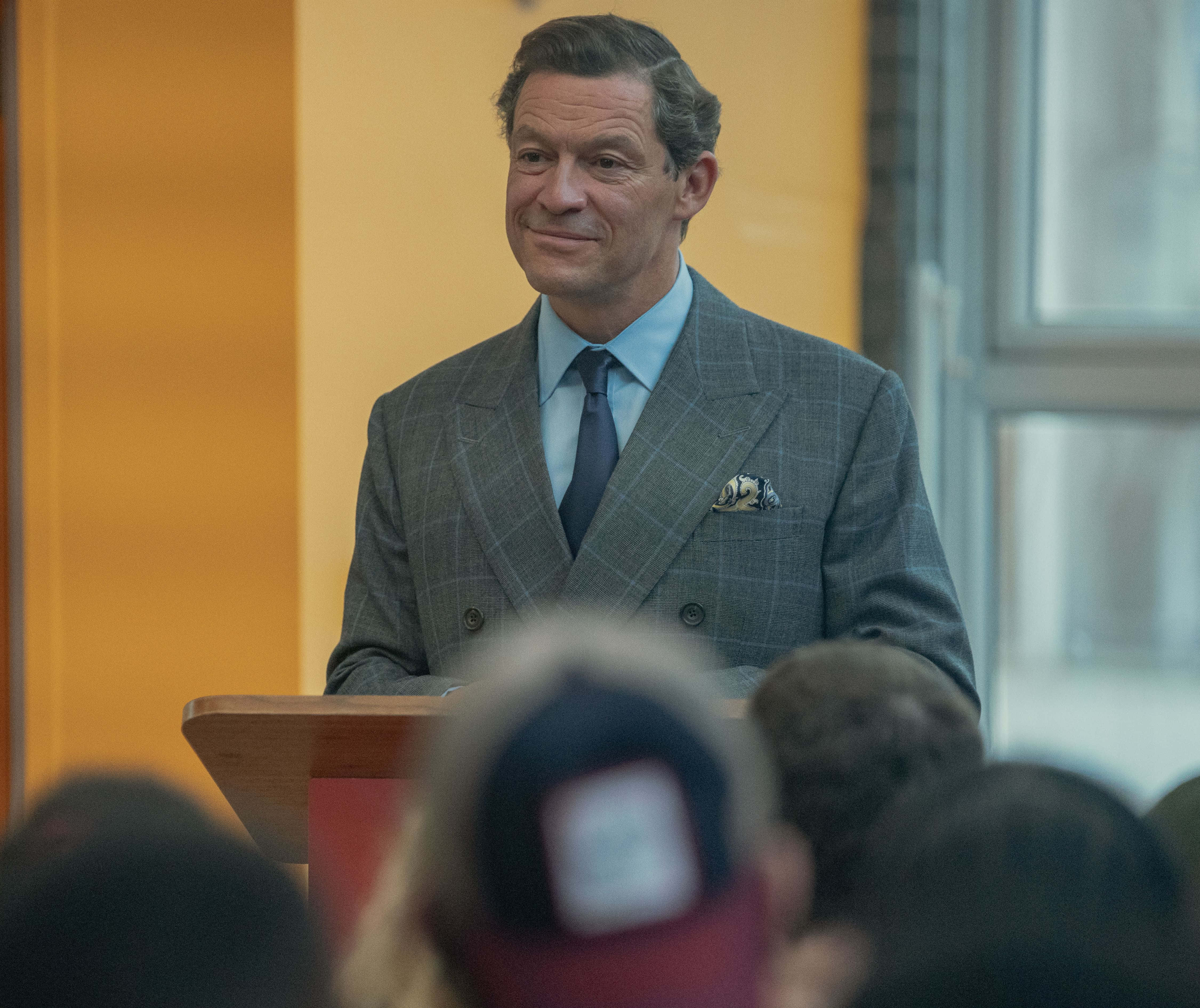 Dominic West 桌布 壁紙 — 一名身穿灰色格紋西裝的男子站在木製講台上，面帶溫暖微笑向觀眾致辭。