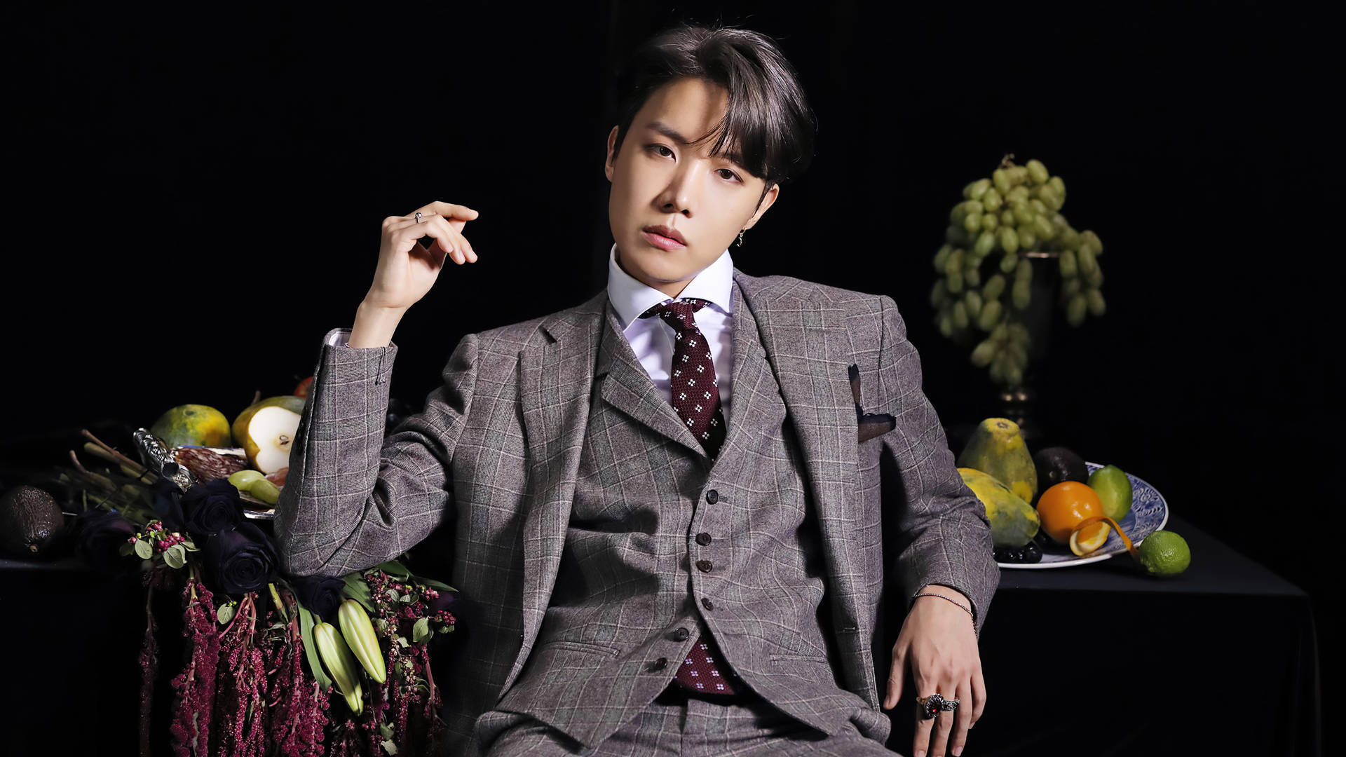 jhope 桌布 壁紙 — 一名身穿灰色格紋西裝的男子自信地坐在黑色背景前，旁邊擺放著色彩鮮豔的水果和花卉靜物。