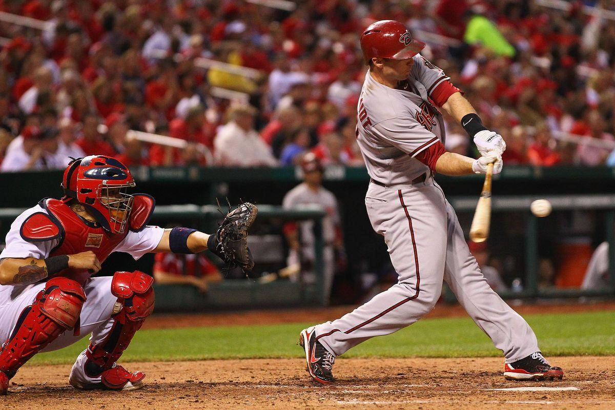 Paul Goldschmidt 桌布 壁紙 — 一名身穿灰色球衣的棒球打者揮棒，而紅色球衣的捕手在球場燈光下蹲在本壘板後方。
