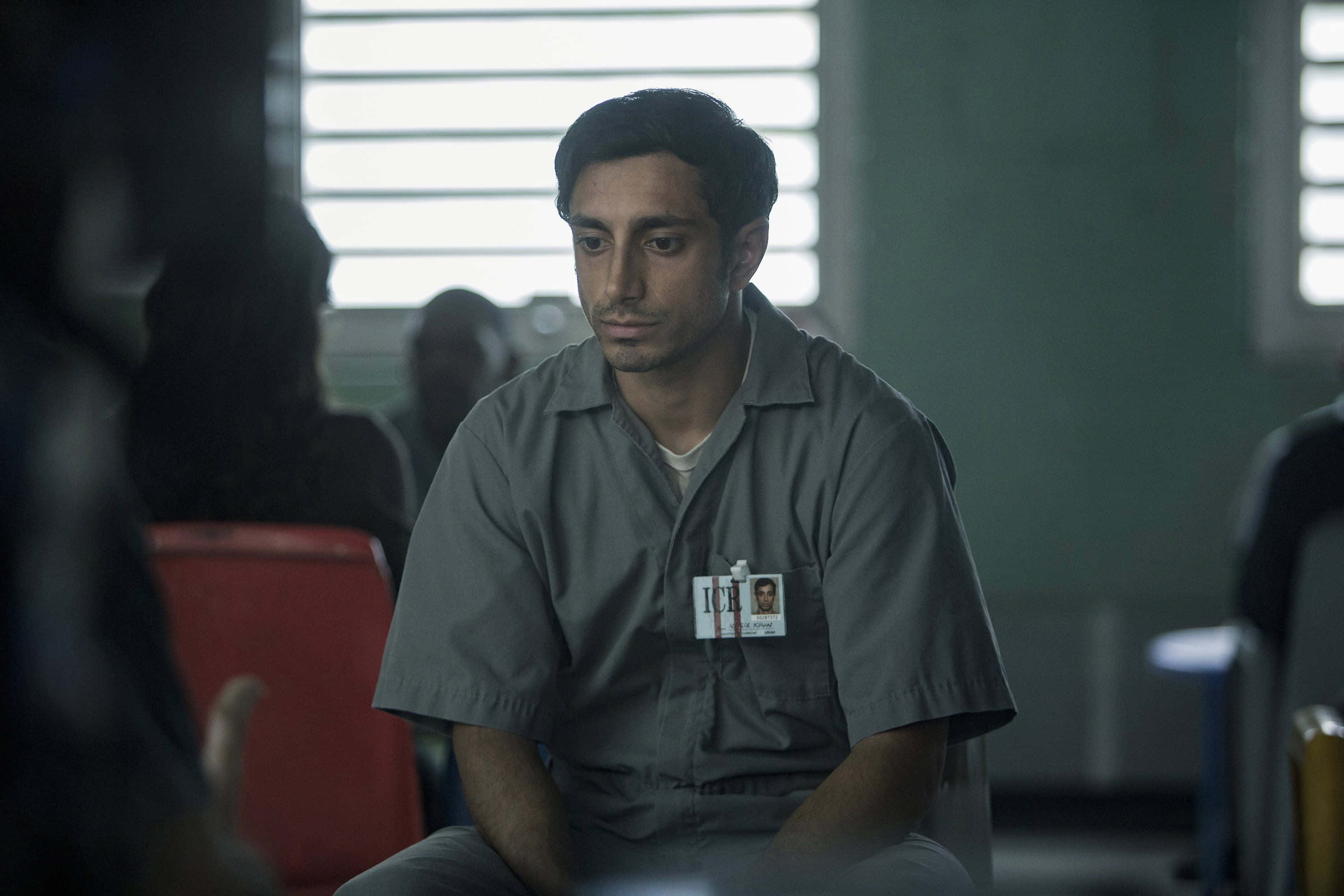 John Turturro 桌布 壁紙 — 一名身穿灰色監獄制服的男人，莊嚴地坐在一個光線昏暗、窗戶有鐵欄的機構房間裡。
