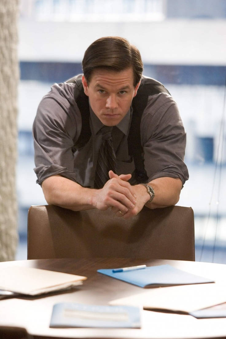 Mark Wahlberg 桌布 壁紙 — 一名身穿灰色襯衫和吊帶褲的男子，在現代辦公室環境中，專注地俯身在一張木桌上。