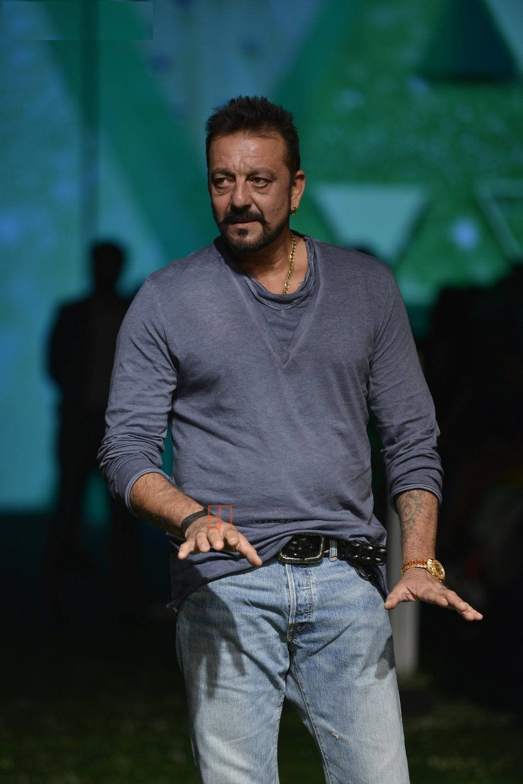 Sanjay 桌布 壁紙 — 一名身穿灰色襯衫和牛仔褲的男子，在綠色燈光背景下張開雙手站立。