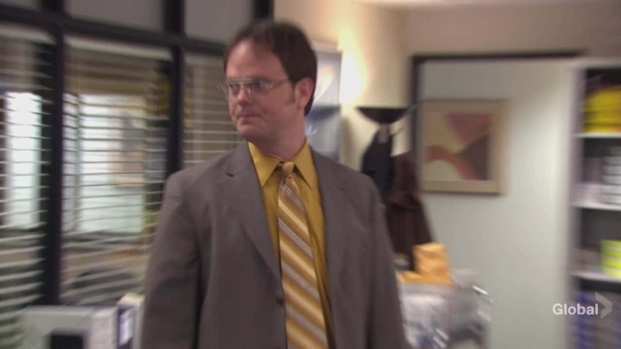 dwight schrute 桌布 壁紙 — 一名身穿灰色西裝、佩戴黃色條紋領帶的男子，走過背景有百葉窗和架子的模糊辦公室。
