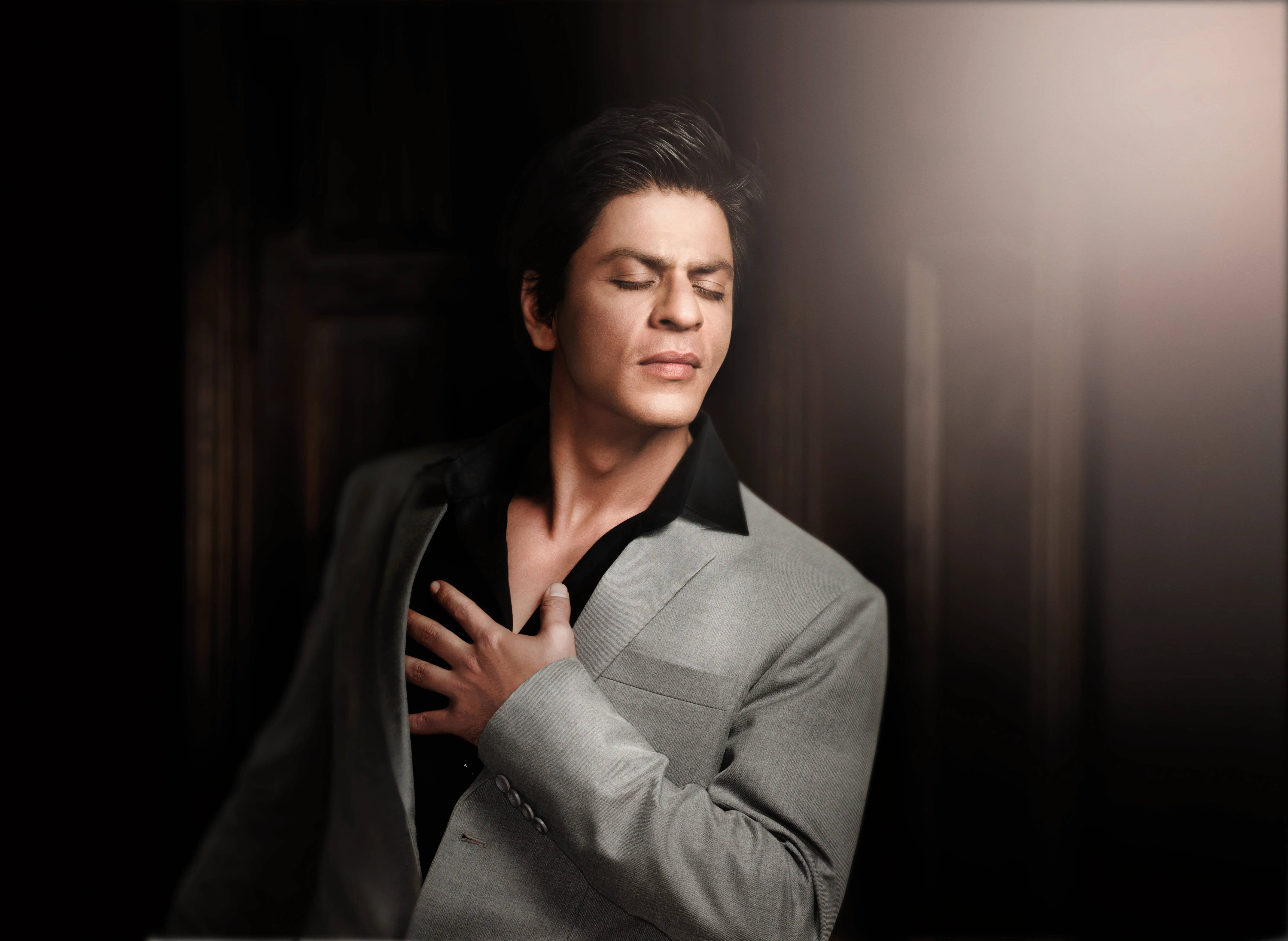 Bollywood HD 桌布 壁紙 — 一名身穿灰色西裝、閉上眼睛的男子，在黑暗柔和的光線背景下輕輕觸碰胸口。