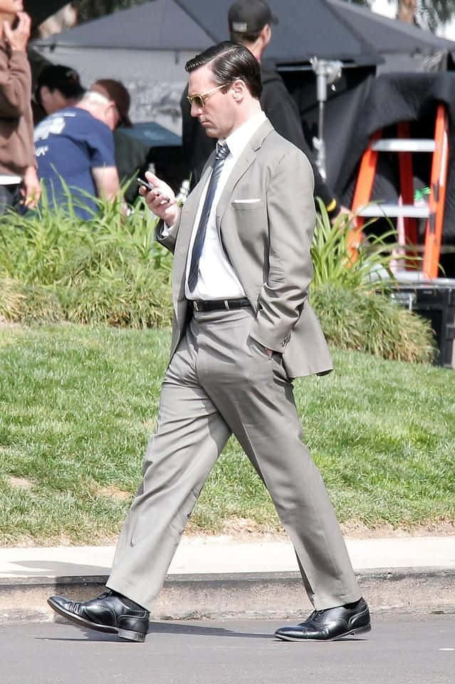Jon Hamm 桌布 壁紙 — 一名身穿灰色西裝和太陽眼鏡的男子，專注地看著手機走在人行道上。
