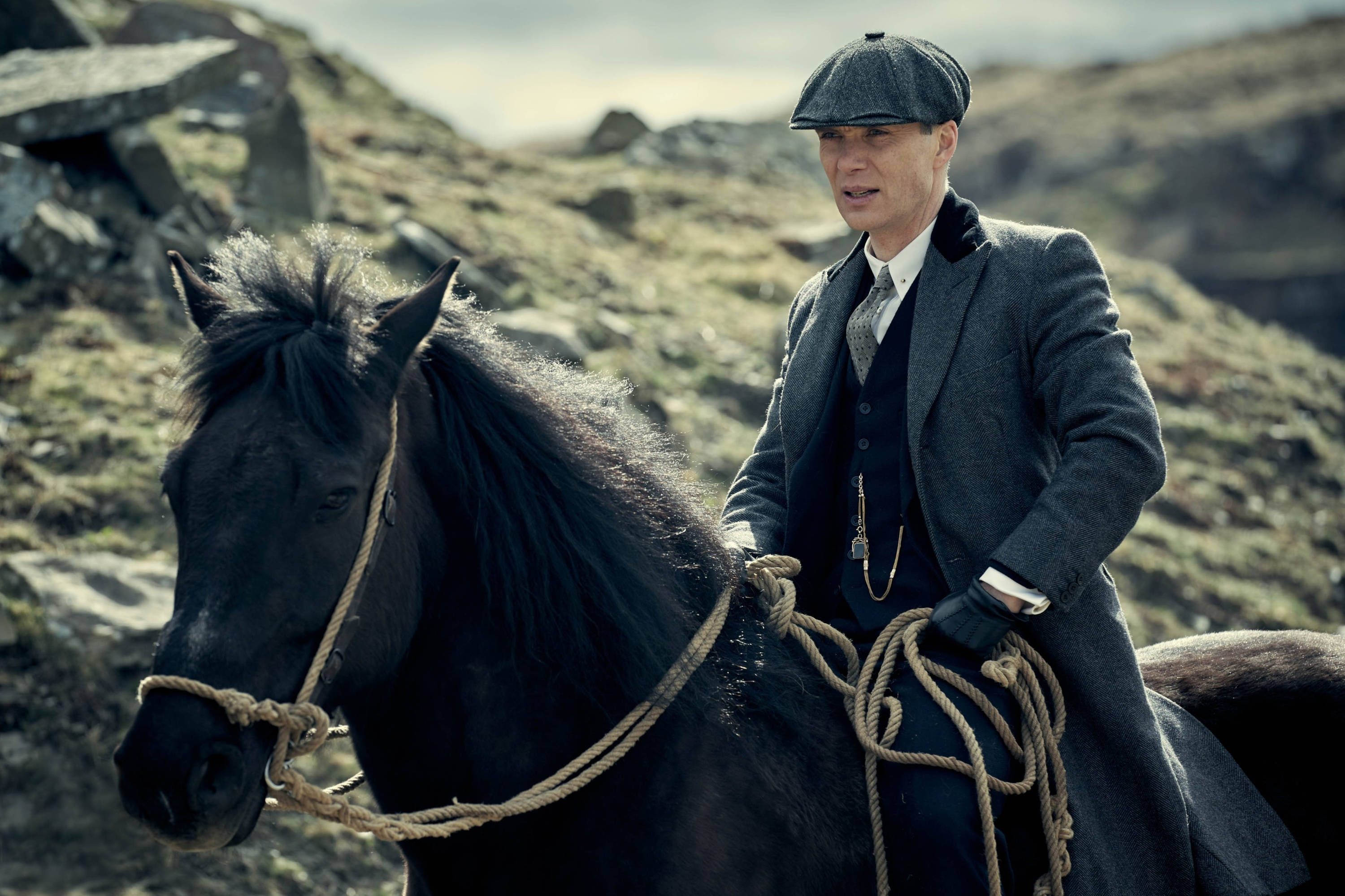 Thomas Shelby 桌布 壁紙 — 一名身穿灰色西裝和扁平帽的男子，在陰天騎著一匹黑馬穿越崎嶇多石的山坡。