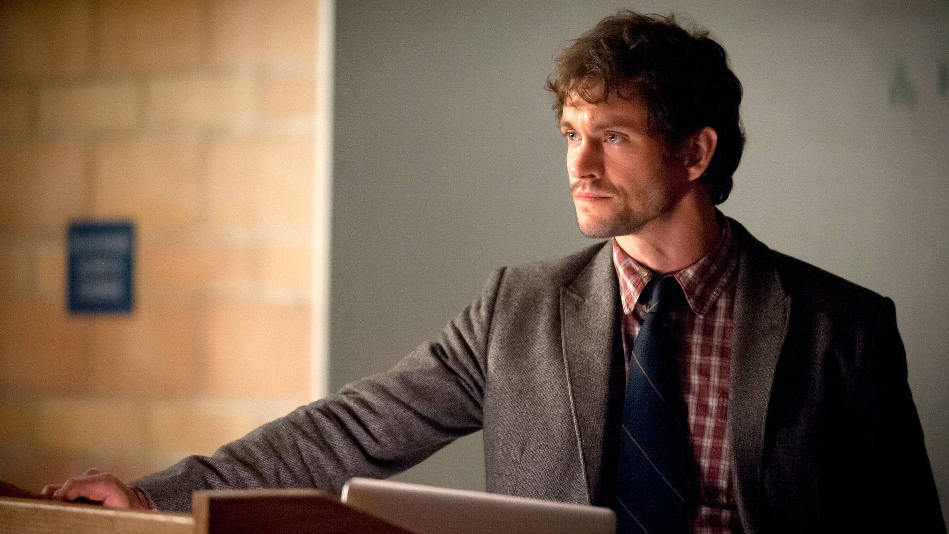 Hugh Dancy 桌布 壁紙 — 一名身穿灰色西裝和格紋襯衫的男子站在一個光線昏暗、磚牆房間的木製講台上。