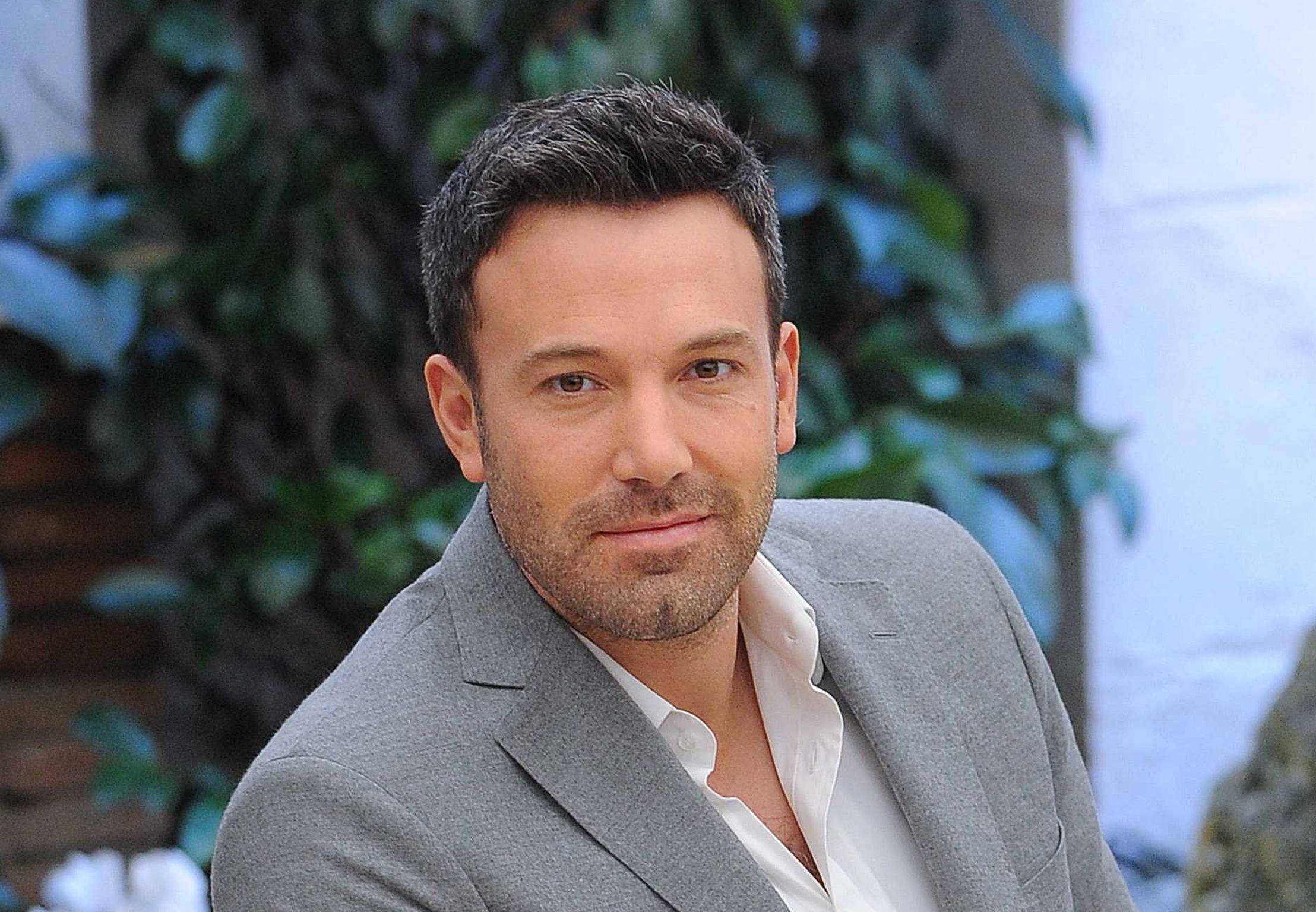 Ben Affleck 桌布 壁紙 — 一名身穿灰色西裝和白色襯衫的男子自信地站在茂密綠色樹葉和白色牆壁的背景前。