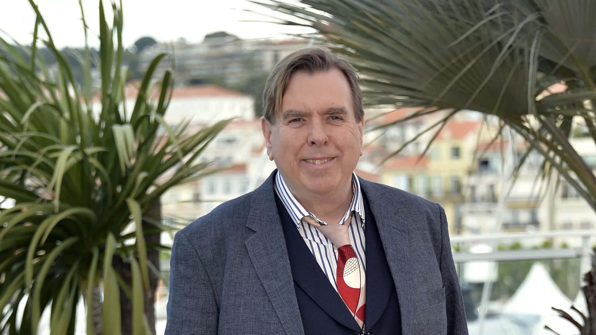 Timothy Spall 桌布 壁紙 — 一名身穿灰色西裝和紅色領帶的男子對著鏡頭微笑，背景是棕櫚樹和沿海小鎮。