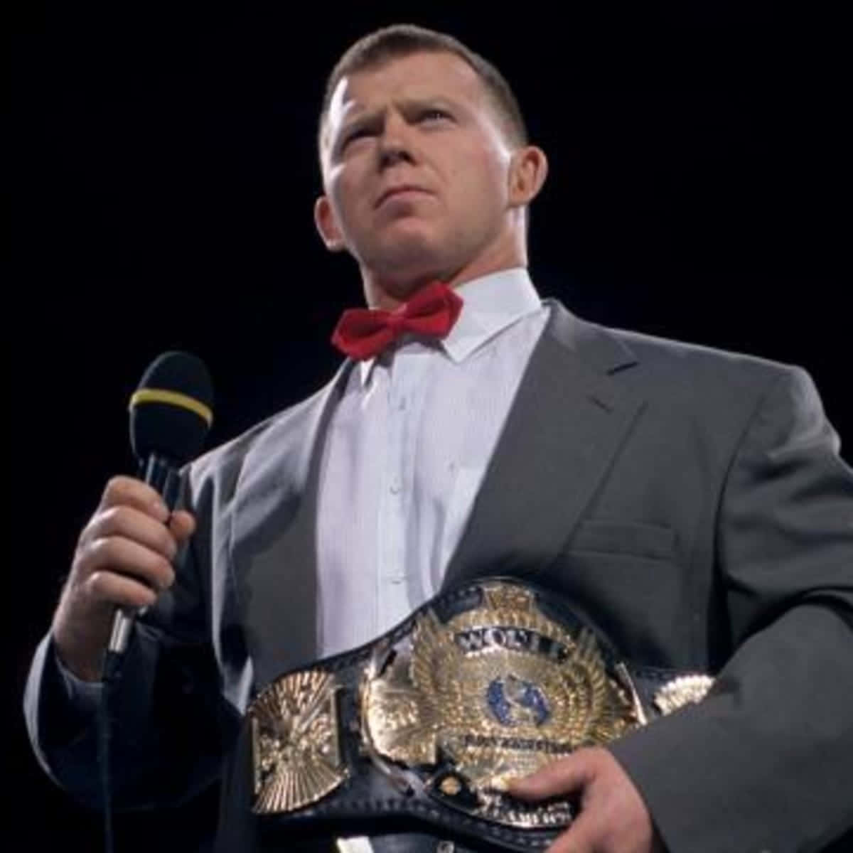 WWE 桌布 壁紙 — 一名身穿灰色西裝和紅色領結的摔角手，在黑色背景前手持冠軍腰帶和麥克風。