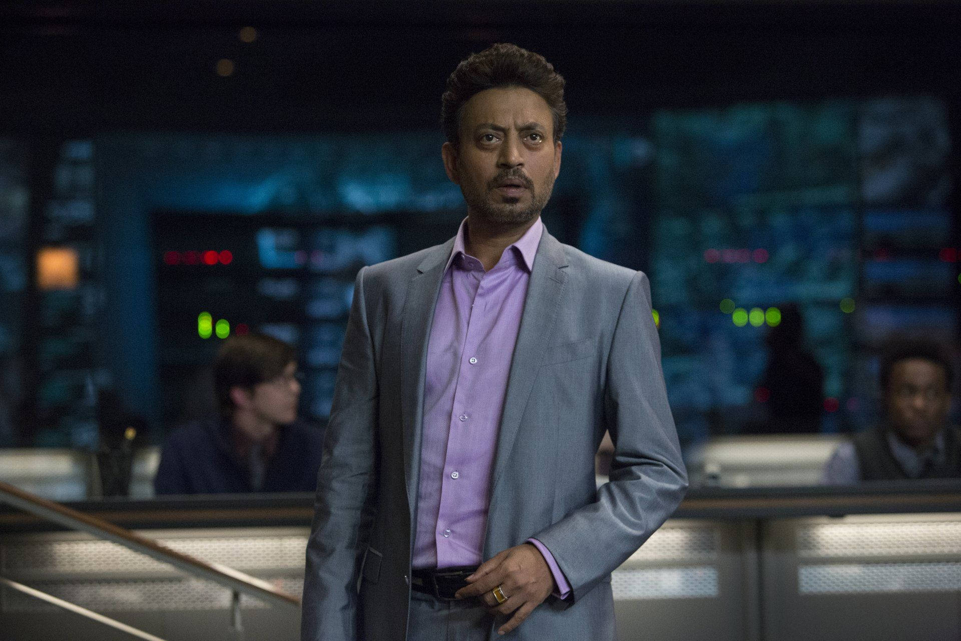 Irrfan Khan 桌布 壁紙 — 一名身穿灰色西裝和紫色襯衫的男子站在高科技控制室中，身後有發光的螢幕和模糊的同事。