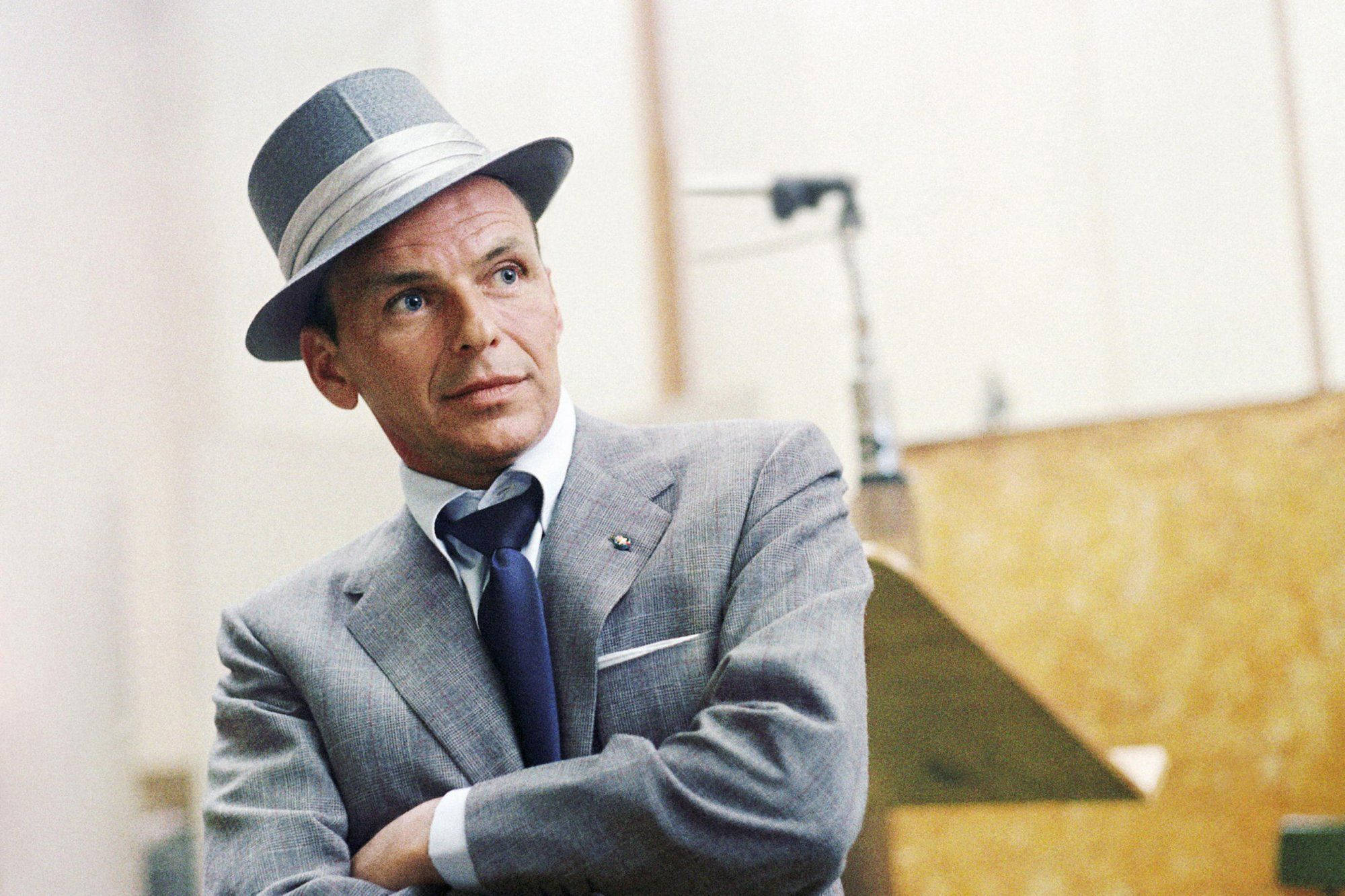 Sinatra 桌布 壁紙 — 一名身穿灰色西裝和軟呢帽的男子雙臂交叉站立，抬頭仰望，處於正式的室內環境中。