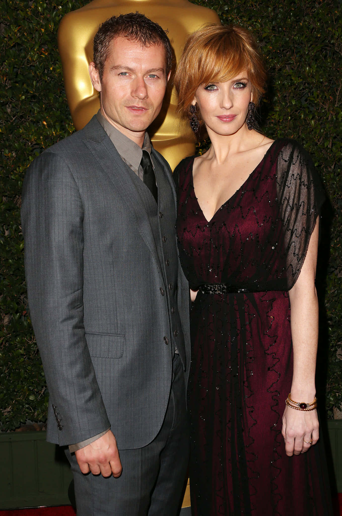 Kelly Reilly 桌布 壁紙 — 一名身穿灰色西裝的男子和一名身穿栗色禮服的女子在頒獎典禮上以金色雕像為背景合影。