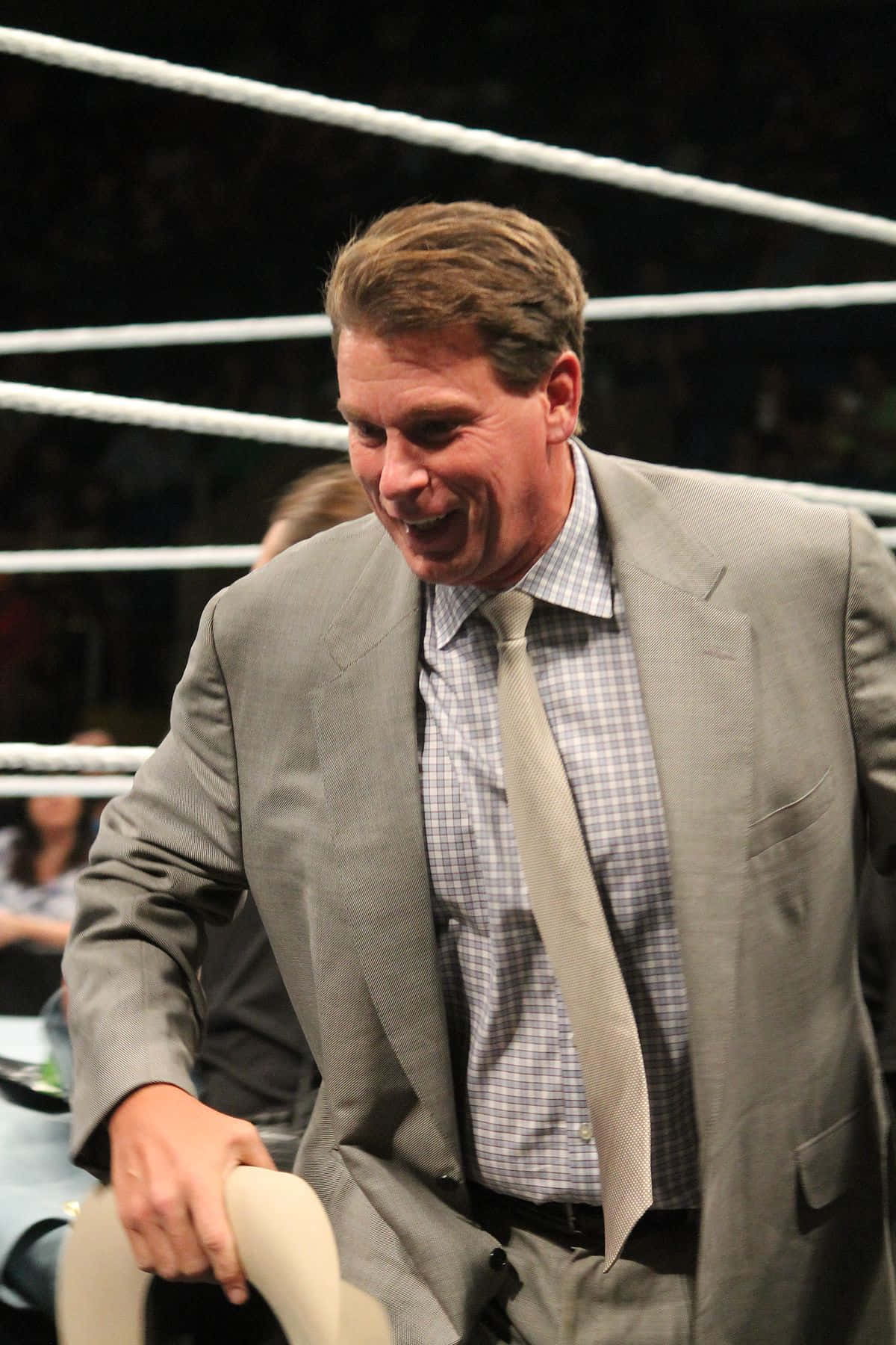 WWE 桌布 壁紙 — 一名身穿灰色西裝的男子在明亮的競技場燈光下，在摔角場內拿著椅子大笑。