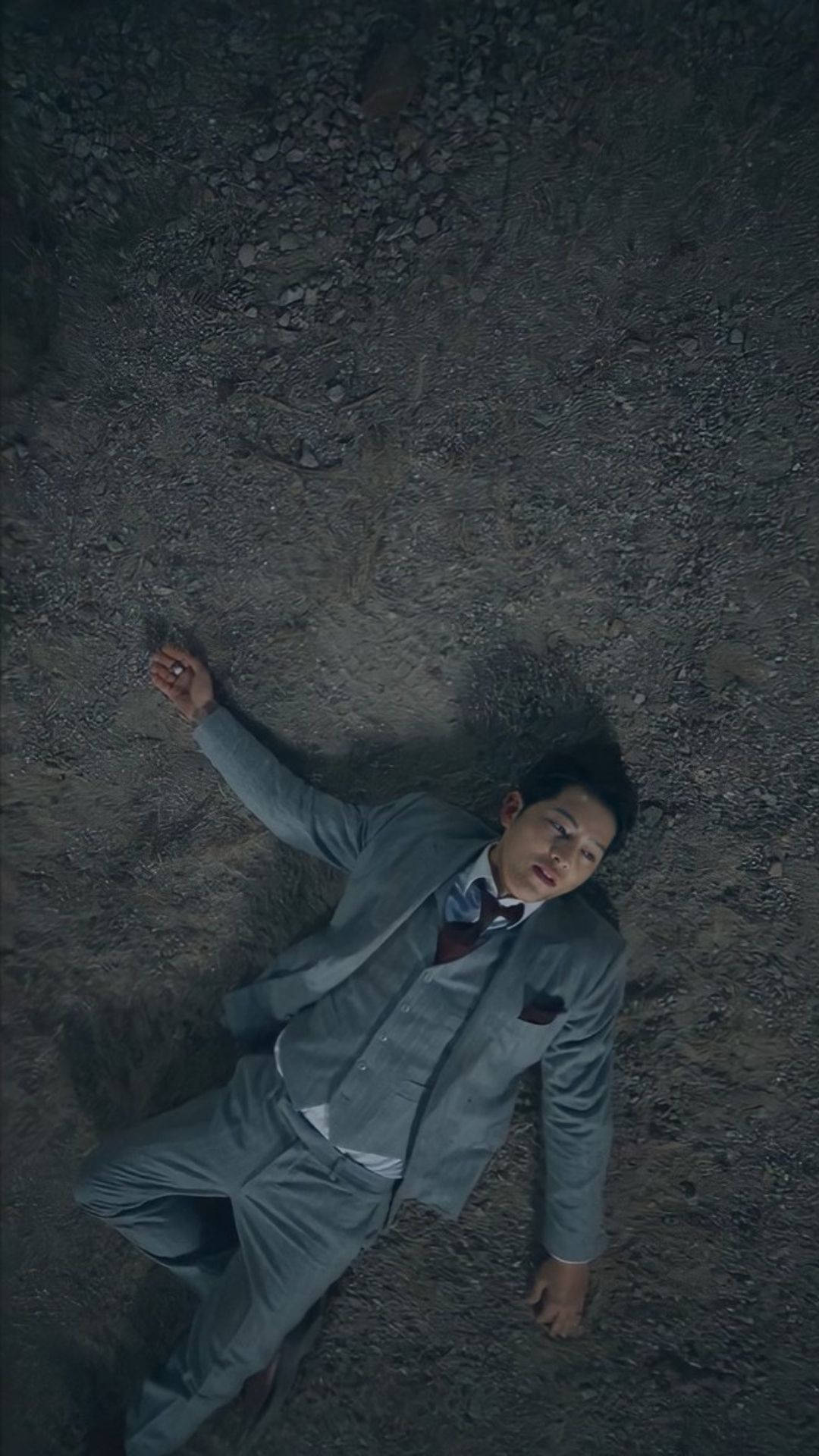 文森佐 桌布 壁紙 — 一名身穿灰色西裝的男子在昏暗、陰鬱的燈光下一動不動地躺在碎石上。
