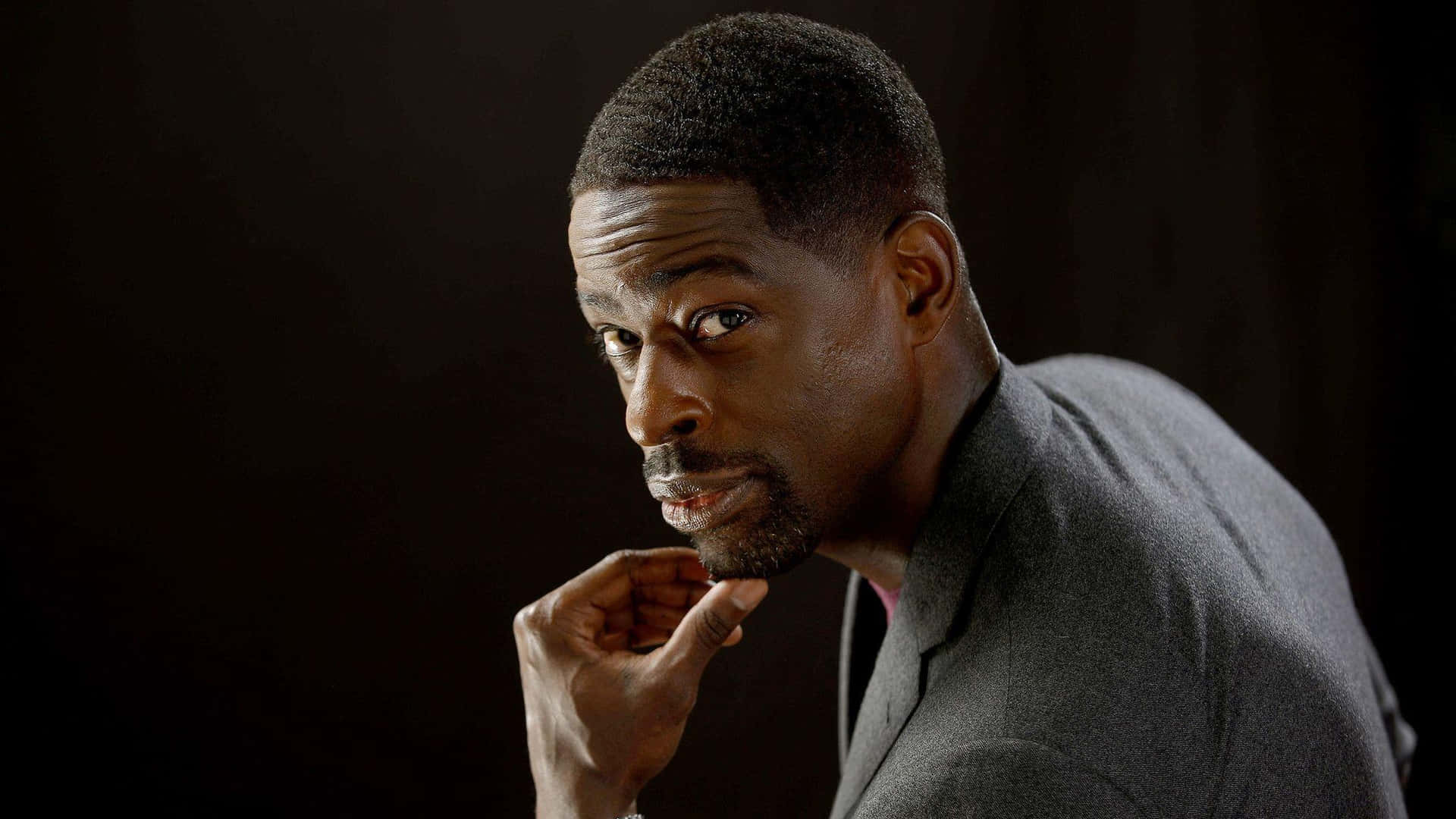 Sterling K Brown 桌布 壁紙 — 一名身穿灰色西裝的男子在深色背景前身體前傾，手靠近下巴，眼神專注而強烈。