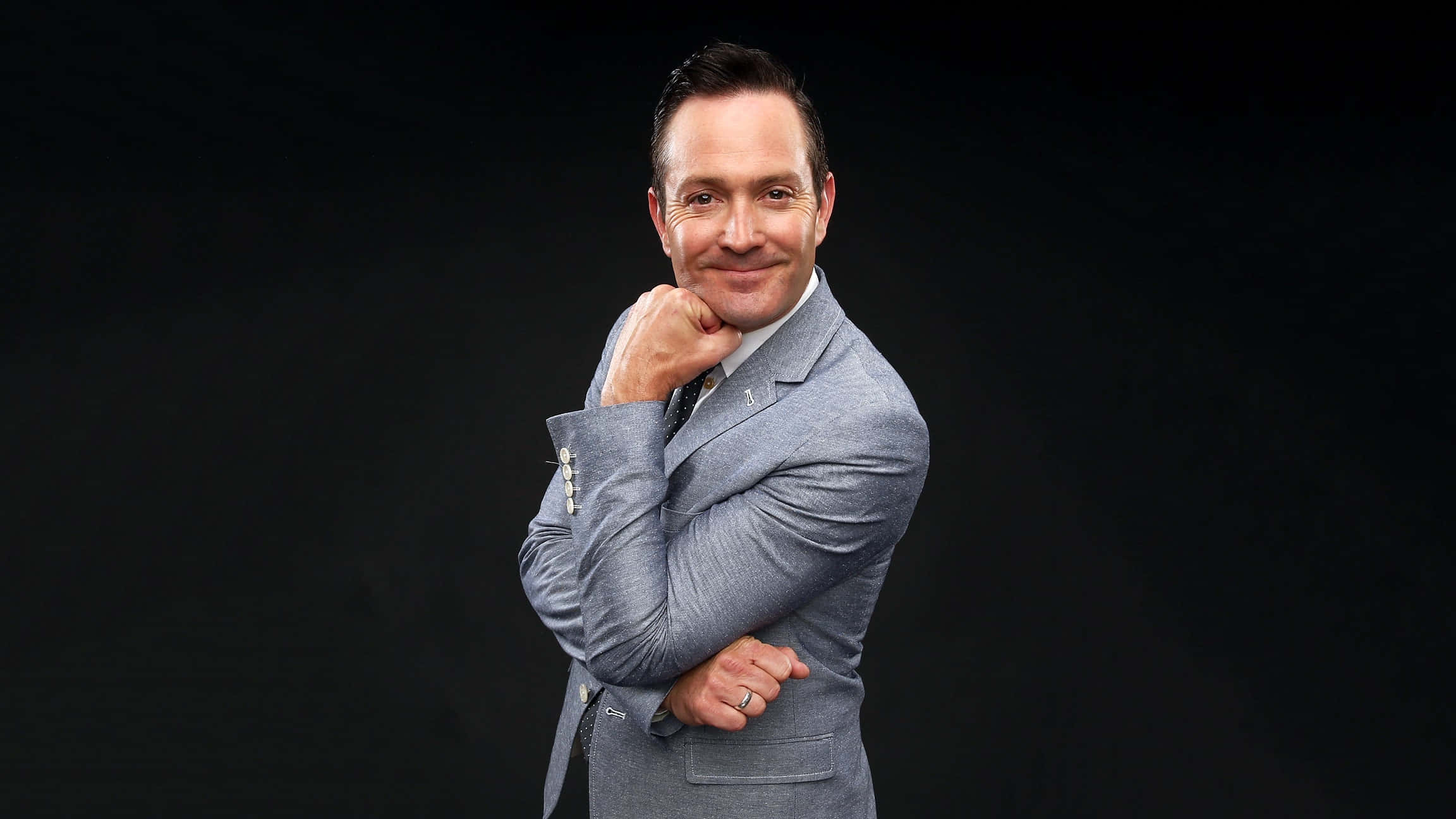 thomas lennon 桌布 壁紙 — 一名身穿灰色西裝的男子在黑色背景前自信地微笑，手托下巴。
