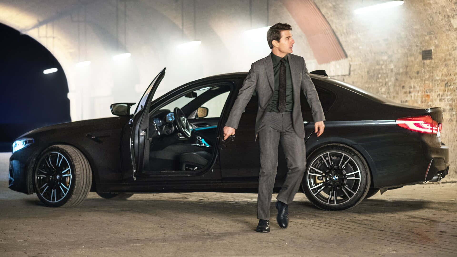 BMW 動圖 桌布 壁紙 — 一名身穿灰色西裝的男子從一輛停在昏暗地下車庫的時尚黑色寶馬轎車中走出，車庫有磚牆和發光的車頭燈。