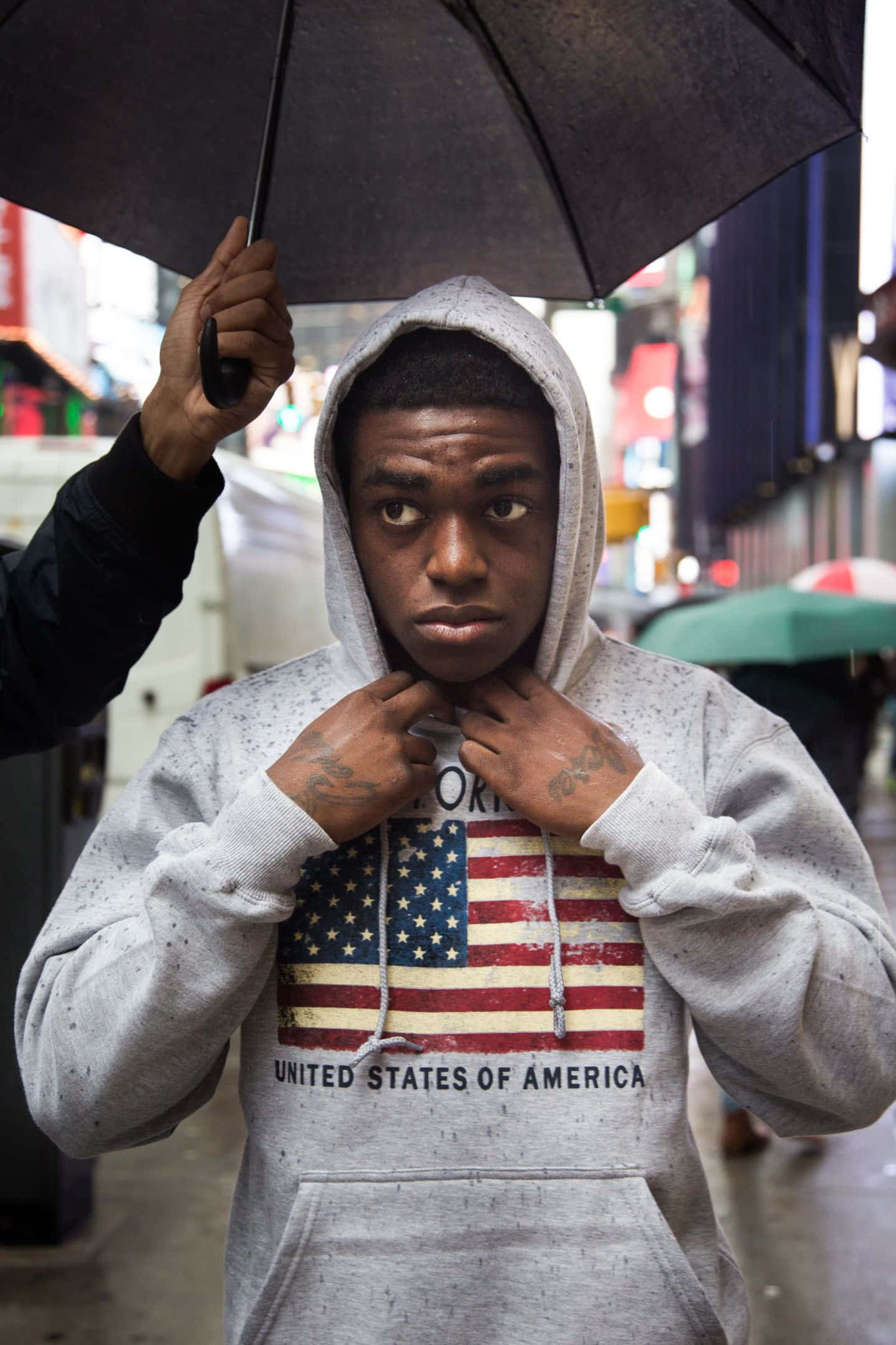 Herbo iPhone 桌布 壁紙 — 一名身穿灰色連帽衫、帶有美國國旗圖案的年輕男子，在雨天的城市街道上，於黑色雨傘下調整衣領。