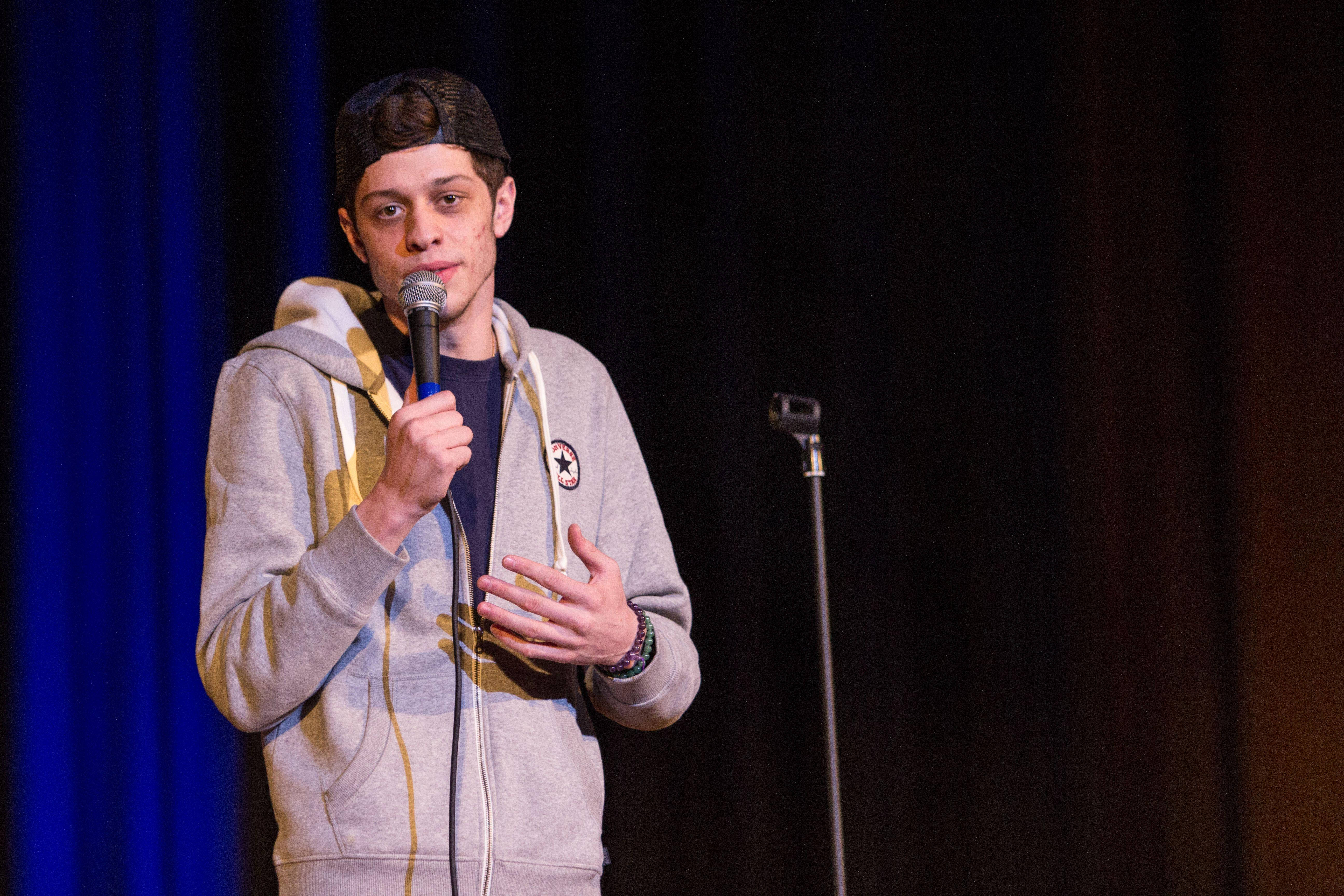 Pete Davidson 桌布 壁紙 — 一名身穿灰色連帽衫和帽子的年輕男子在舞台上拿著麥克風，身後有藍色窗簾。