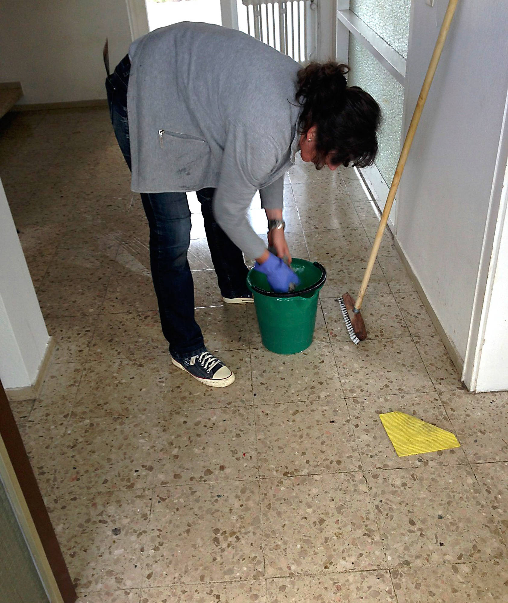 Cleaning 桌布 壁紙 — 一名身穿灰色連帽衫和藍色手套的人正在用拖把清潔有斑點的瓷磚地板，旁邊有一個綠色水桶和掃帚。