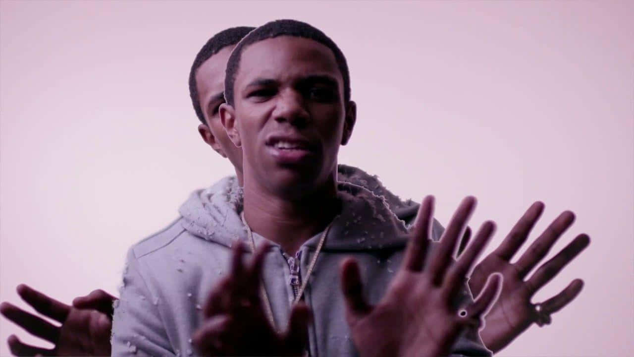 A Boogie Wit Da Hoodie 桌布 壁紙 — 一名身穿灰色連帽衫的男子在柔和的粉紅色背景前用雙手做手勢，帶有微妙的雙重曝光效果。