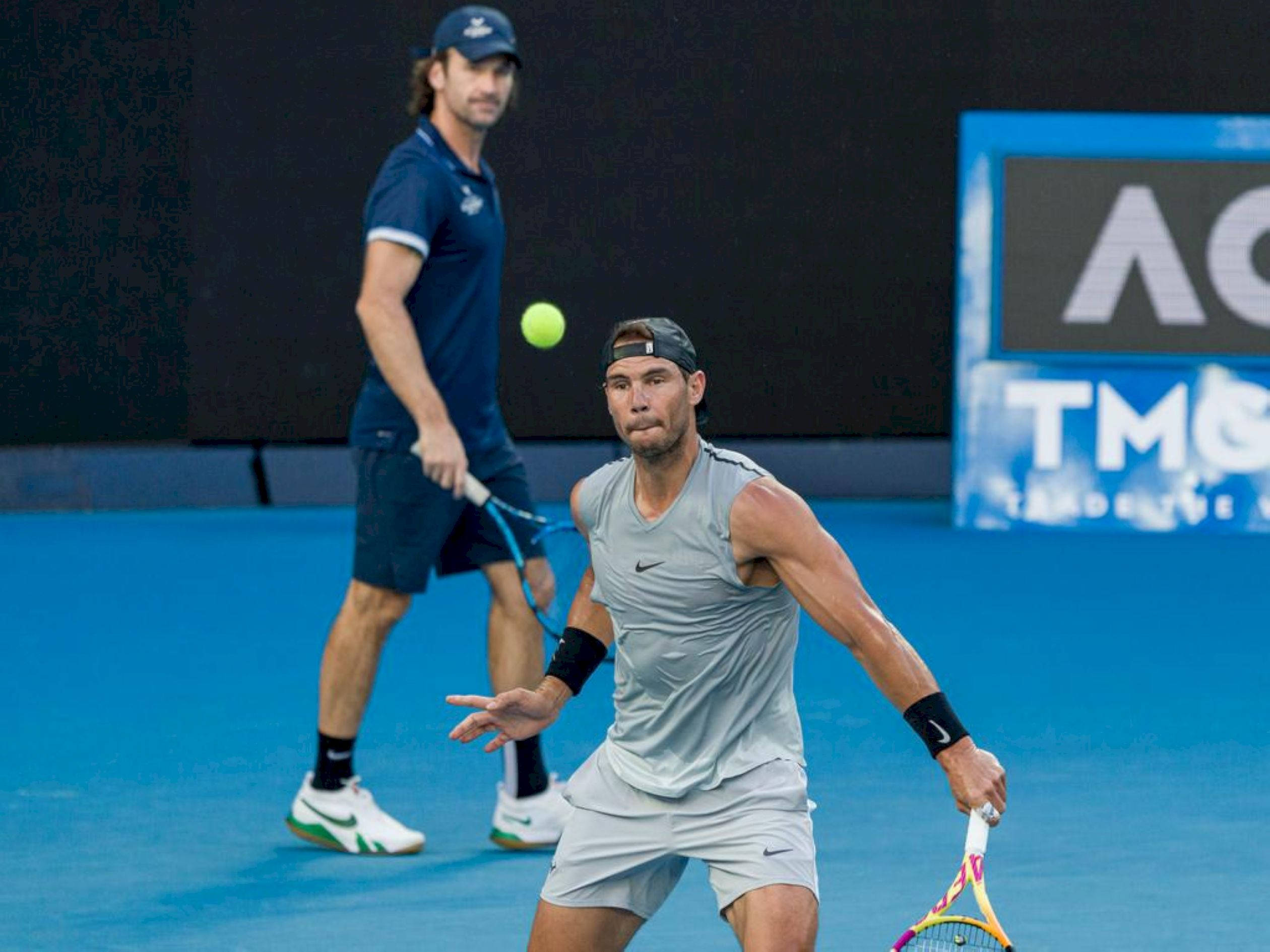 Tennis 桌布 壁紙 — 一名身穿灰色運動服的男網球選手在充滿活力的藍色球場上向前猛衝，擊打一個亮黃色的網球，背景有另一名選手。