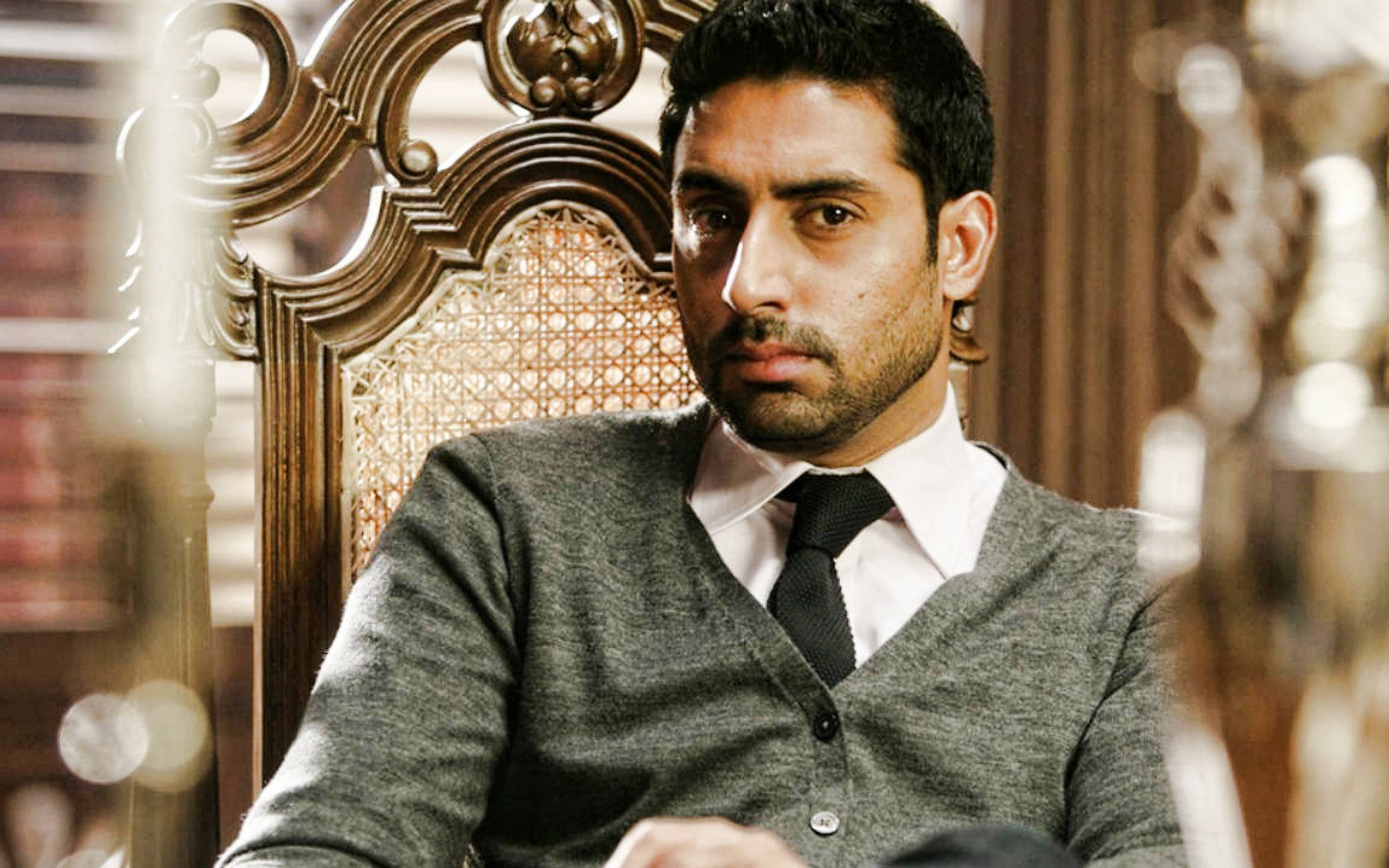 abhishek bachchan 桌布 壁紙 — 一名身穿灰色開襟毛衣、打著領帶的男子，端坐在一張華麗的木椅上，背景是模糊的書架。