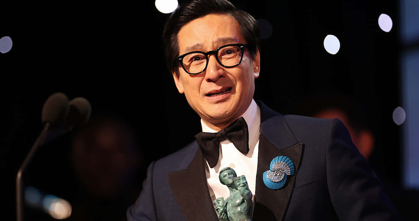 Ke Huy Quan 桌布 壁紙 — 一名身穿燕尾服、戴眼鏡和領結的男子，在燈光昏暗、背後有散景燈光的舞台上手持獎項。