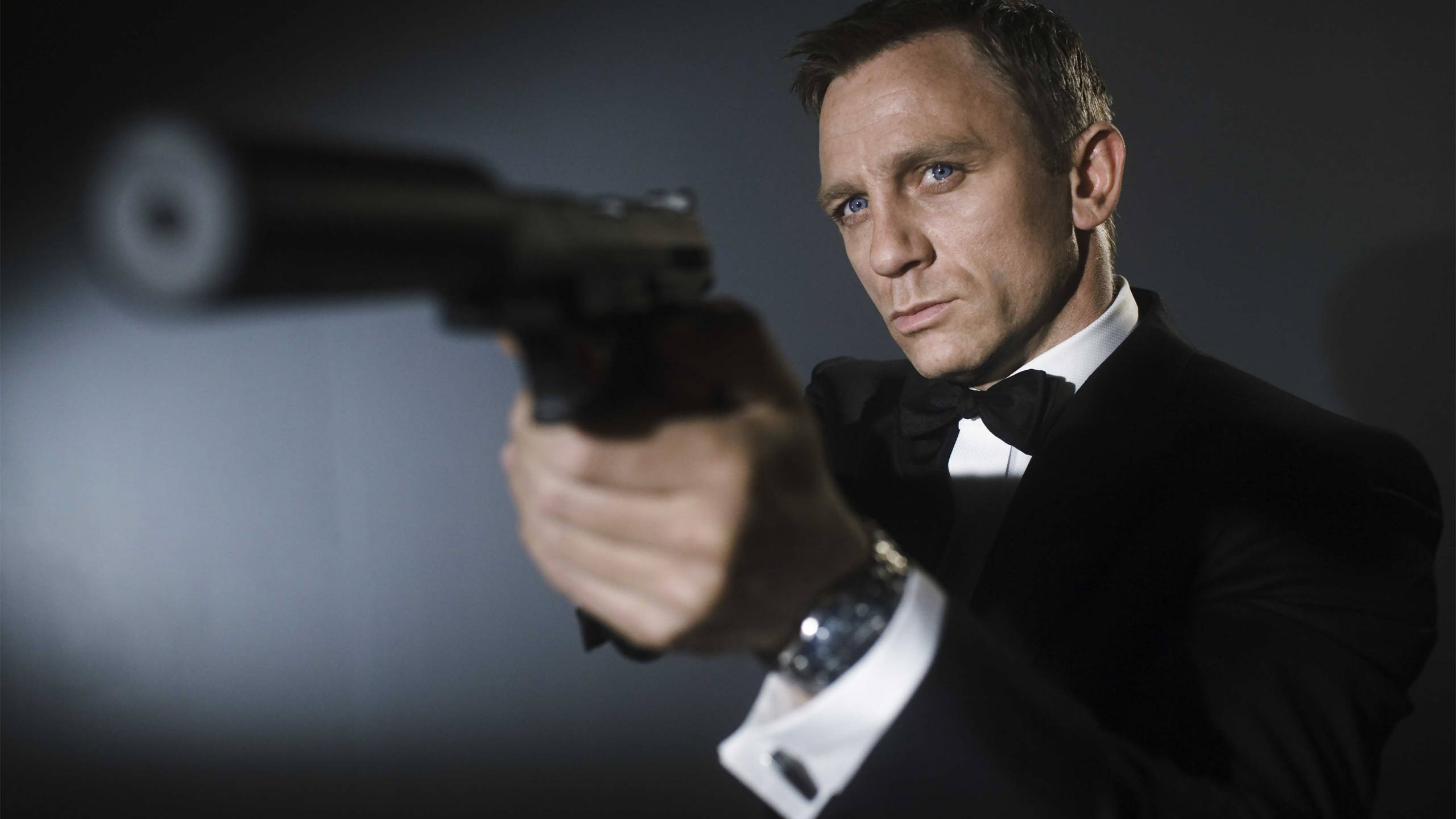 Daniel Craig 桌布 壁紙 — 一名身穿燕尾服的男人，在深色漸層背景前，專注地瞄準一把消音手槍。