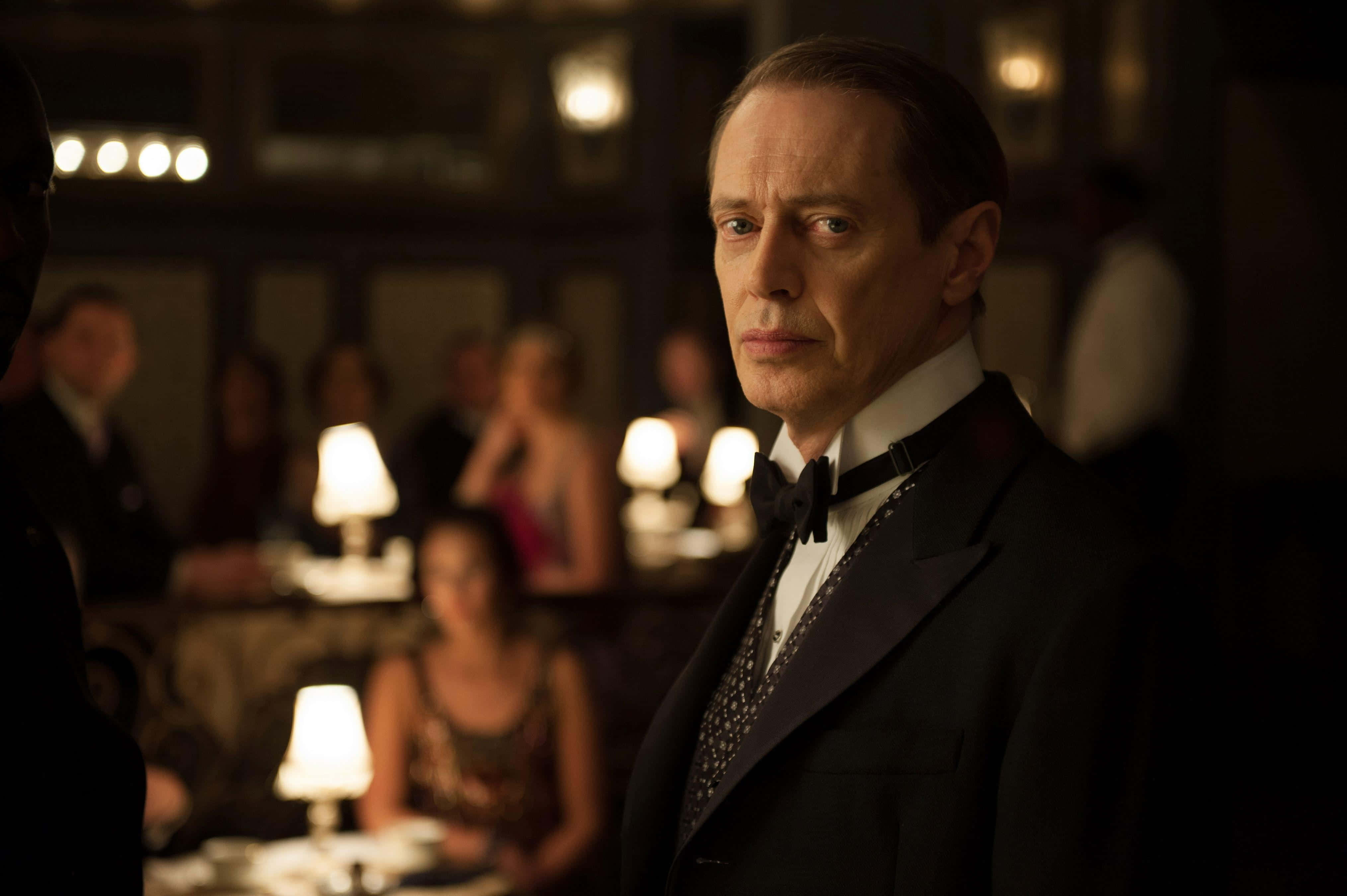 steve buscemi 桌布 壁紙 — 一名身穿燕尾服的男人，在燈光昏暗、氣氛溫暖的高級餐廳中莊嚴地站立，背景有模糊的顧客。