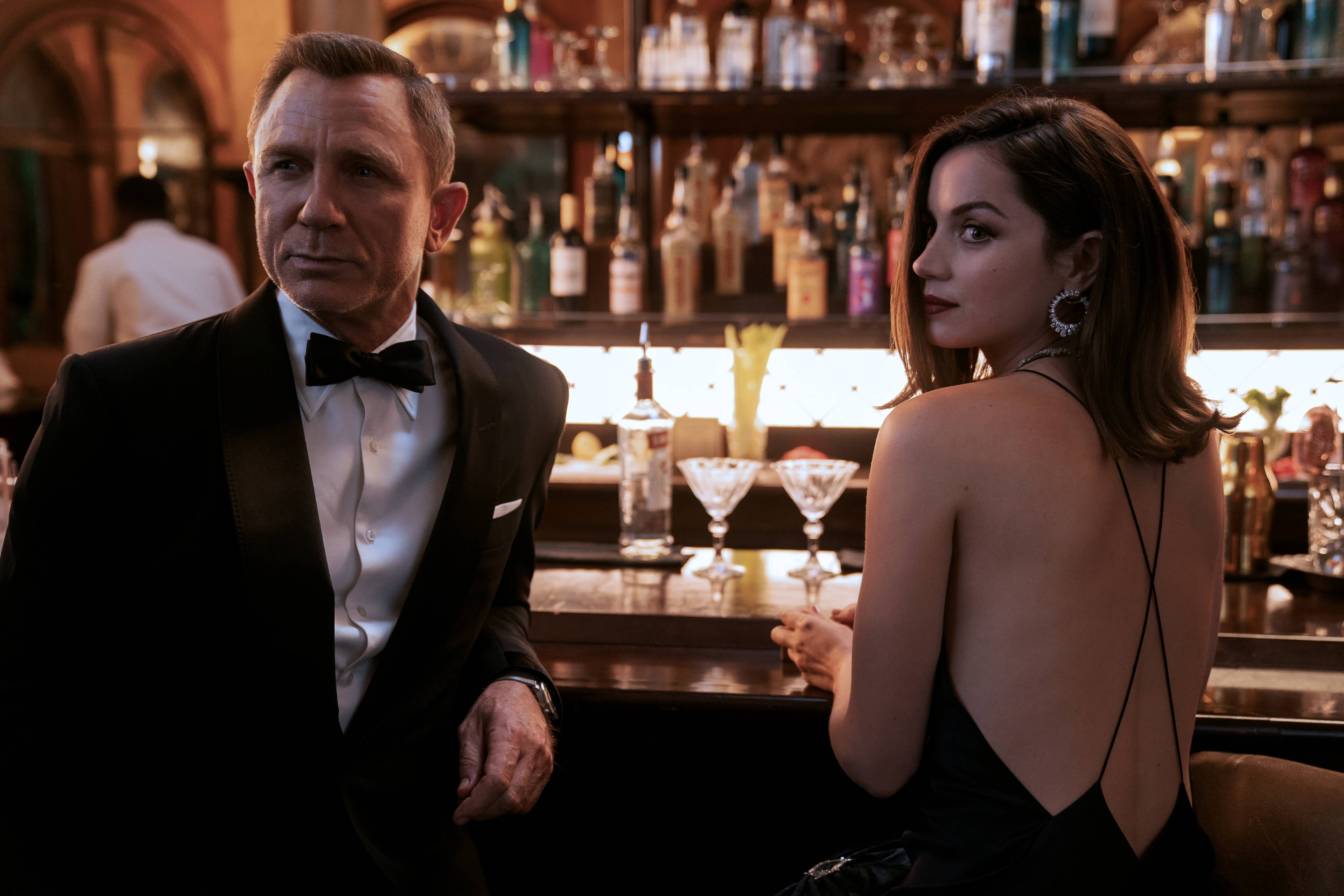 Daniel Craig 桌布 壁紙 — 一名身穿燕尾服的男士和一名身穿黑色連衣裙的女士坐在燈光昏暗的酒吧裡，身後有擺滿酒瓶的架子。