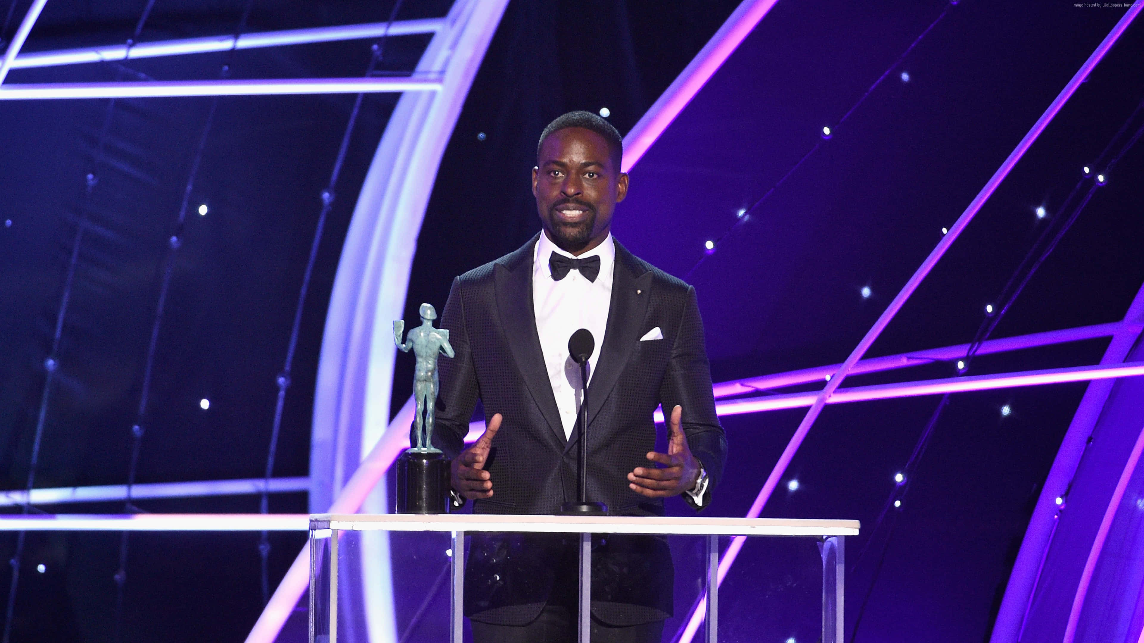 Sterling K Brown 桌布 壁紙 — 一名身穿燕尾服的男士站在講台上，在充滿活力的紫色舞台燈光下手持獎項。