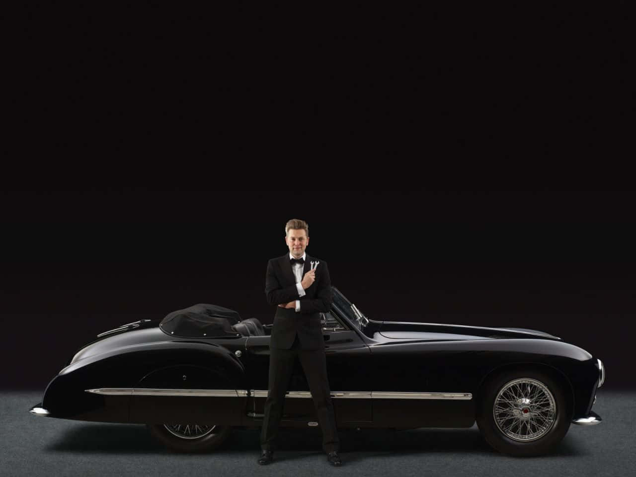 Black And White Vintage Car 桌布 壁紙 — 一名身穿燕尾服的男士自信地站在一輛時尚的黑色復古敞篷車旁，背景是深色攝影棚。