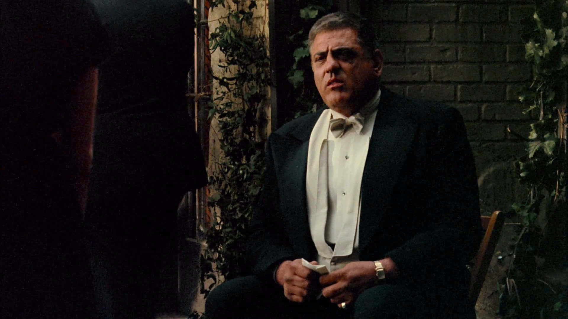 The Godfather 桌布 壁紙 — 一名身穿燕尾服的男士莊嚴地坐在椅子上，背靠長滿常春藤的磚牆，表情嚴肅地向上望。