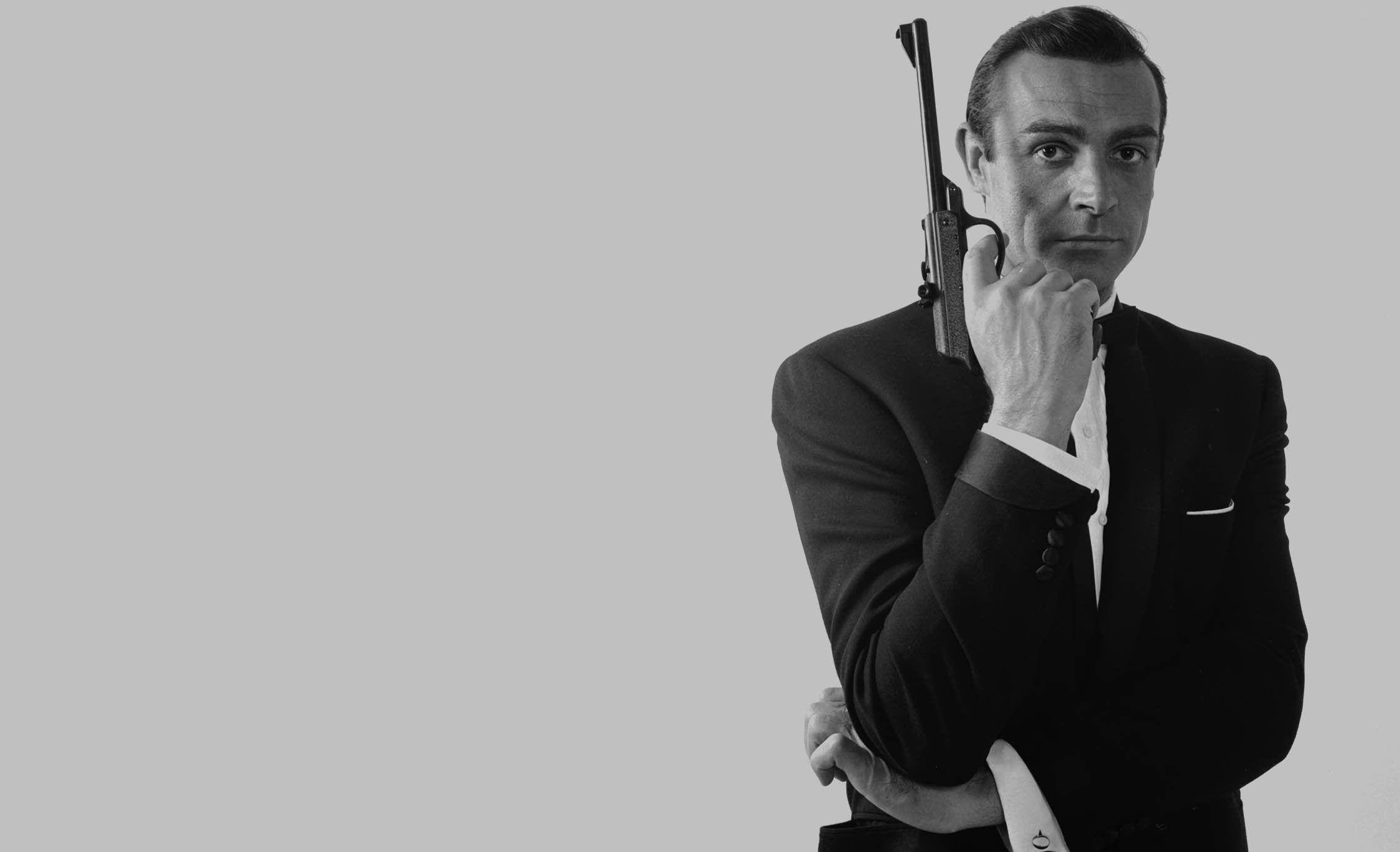 Sean Connery 桌布 壁紙 — 一名身穿燕尾服的男子在純灰色背景下，以黑白鮮明的肖像照形式，將手槍抵住自己的太陽穴。