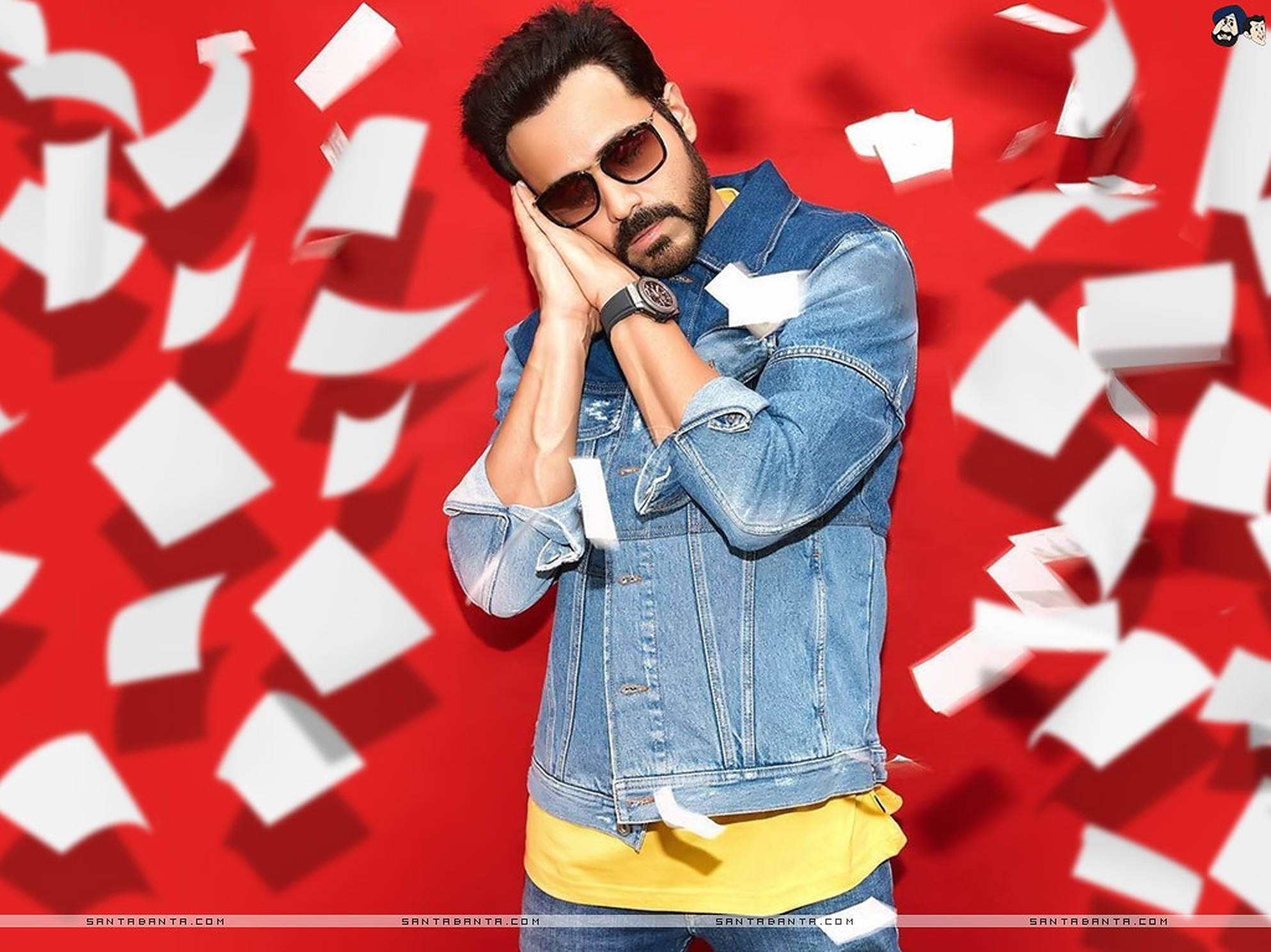 Emraan Hashmi 桌布 壁紙 — 一名身穿牛仔外套和太陽眼鏡的男子站在鮮紅色的背景前，白色紙屑在他周圍旋轉。