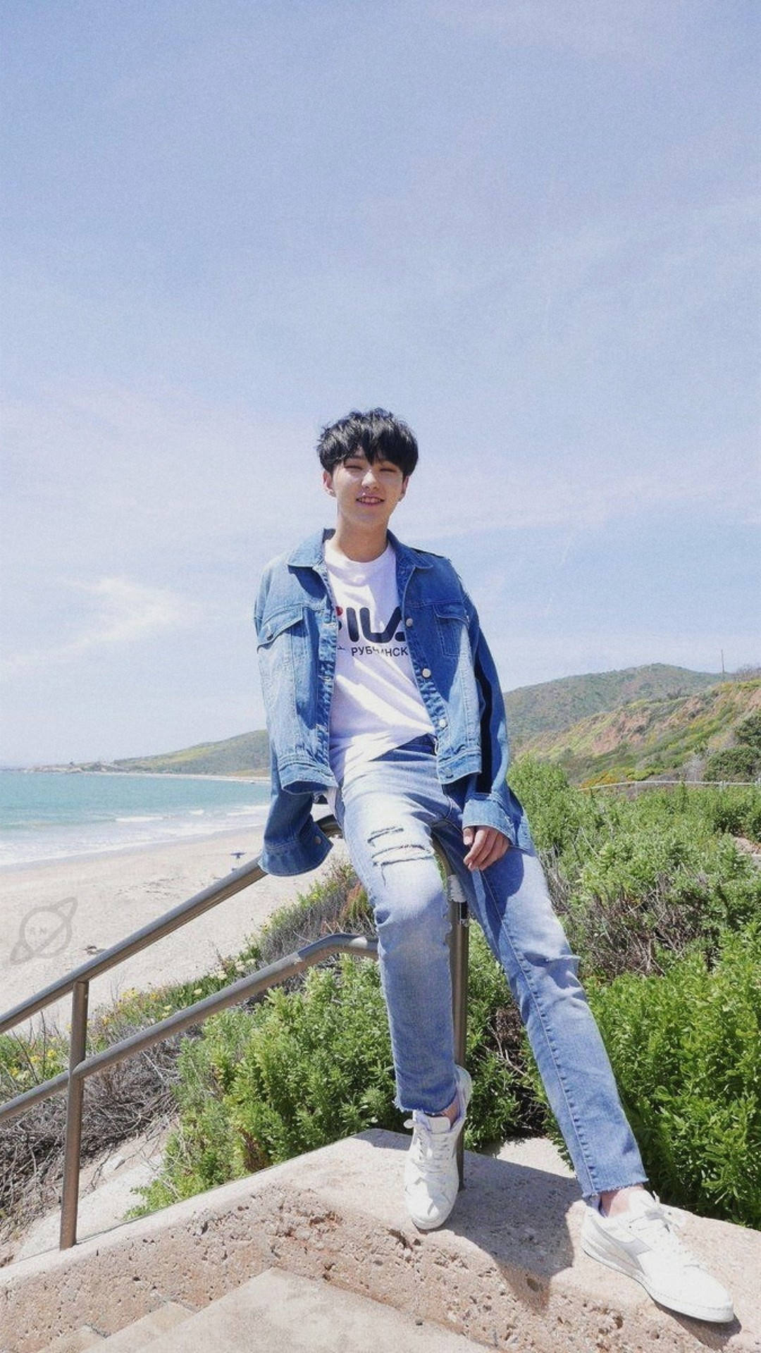 Hoshi 桌布 壁紙 — 一名身穿牛仔外套和破洞牛仔褲的年輕男子，隨意地坐在石階上，俯瞰著陽光普照的海灘，碧綠的海水和翠綠的山丘，天空清澈湛藍。