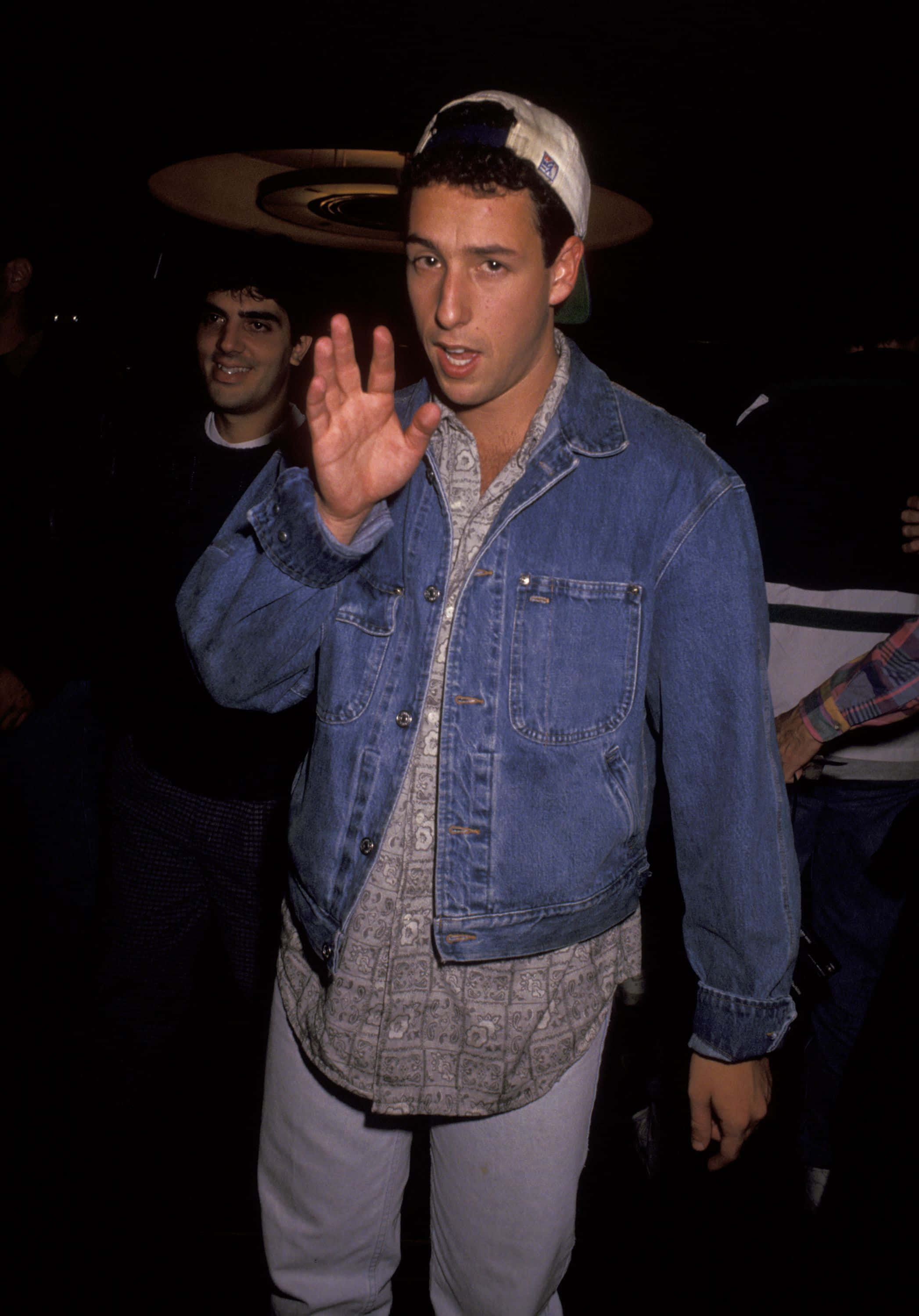 Adam Sandler 桌布 壁紙 — 一名身穿牛仔夾克、戴著反戴帽子的男子在昏暗的夜店燈光下向鏡頭揮手。