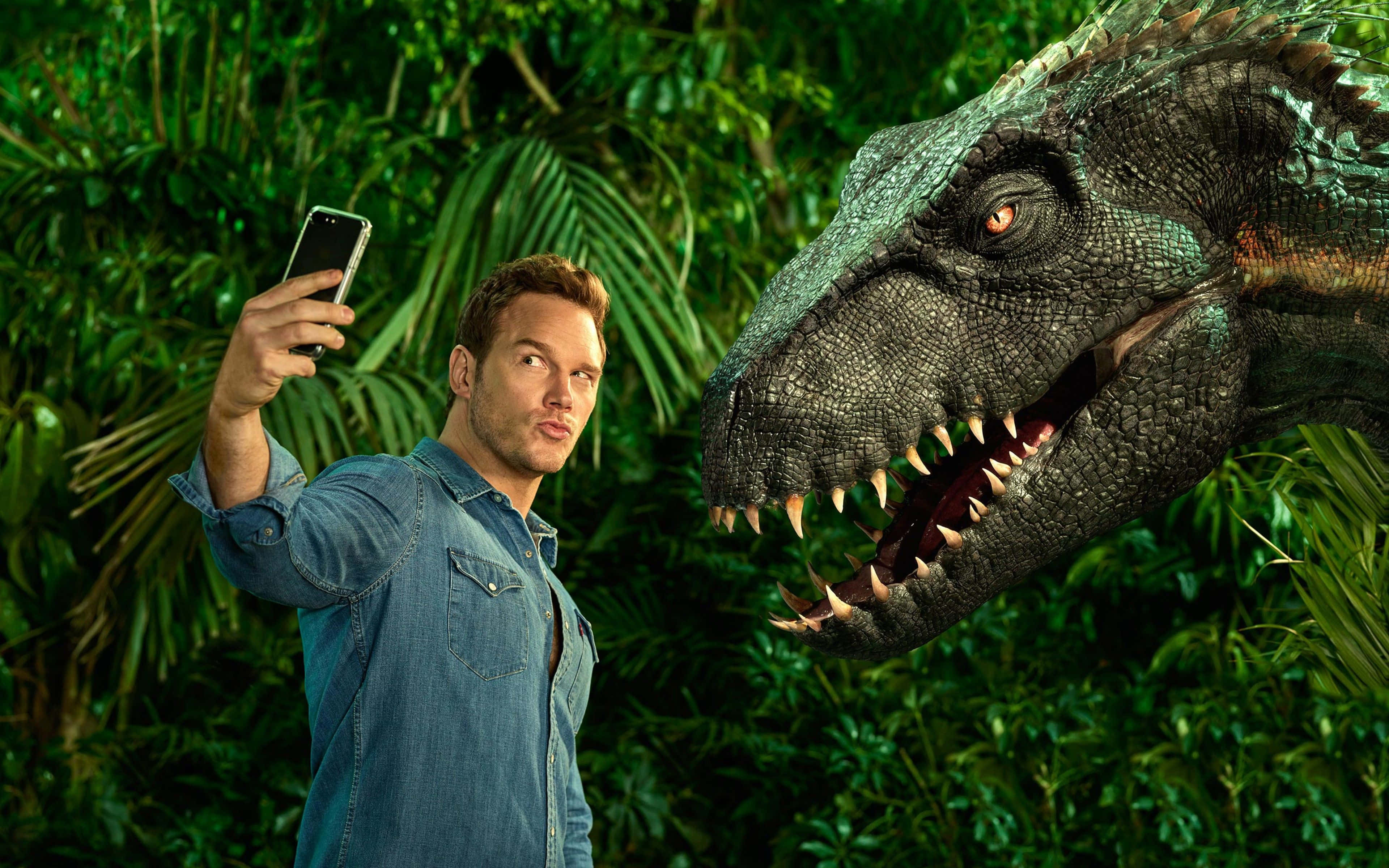 Jurassic World 桌布 壁紙 — 一名身穿牛仔衫的男子在茂密的綠色叢林中與一隻咆哮的恐龍自拍。