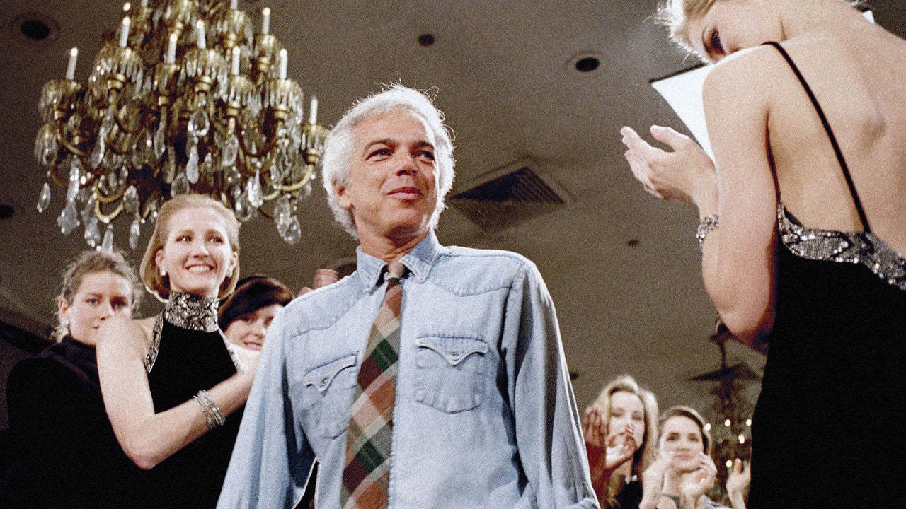ralph lauren 桌布 壁紙 — 一名身穿牛仔襯衫和格子領帶的男子，在一個華麗的室內環境中，周圍有吊燈和衣著優雅的女士們，他溫暖地微笑著。