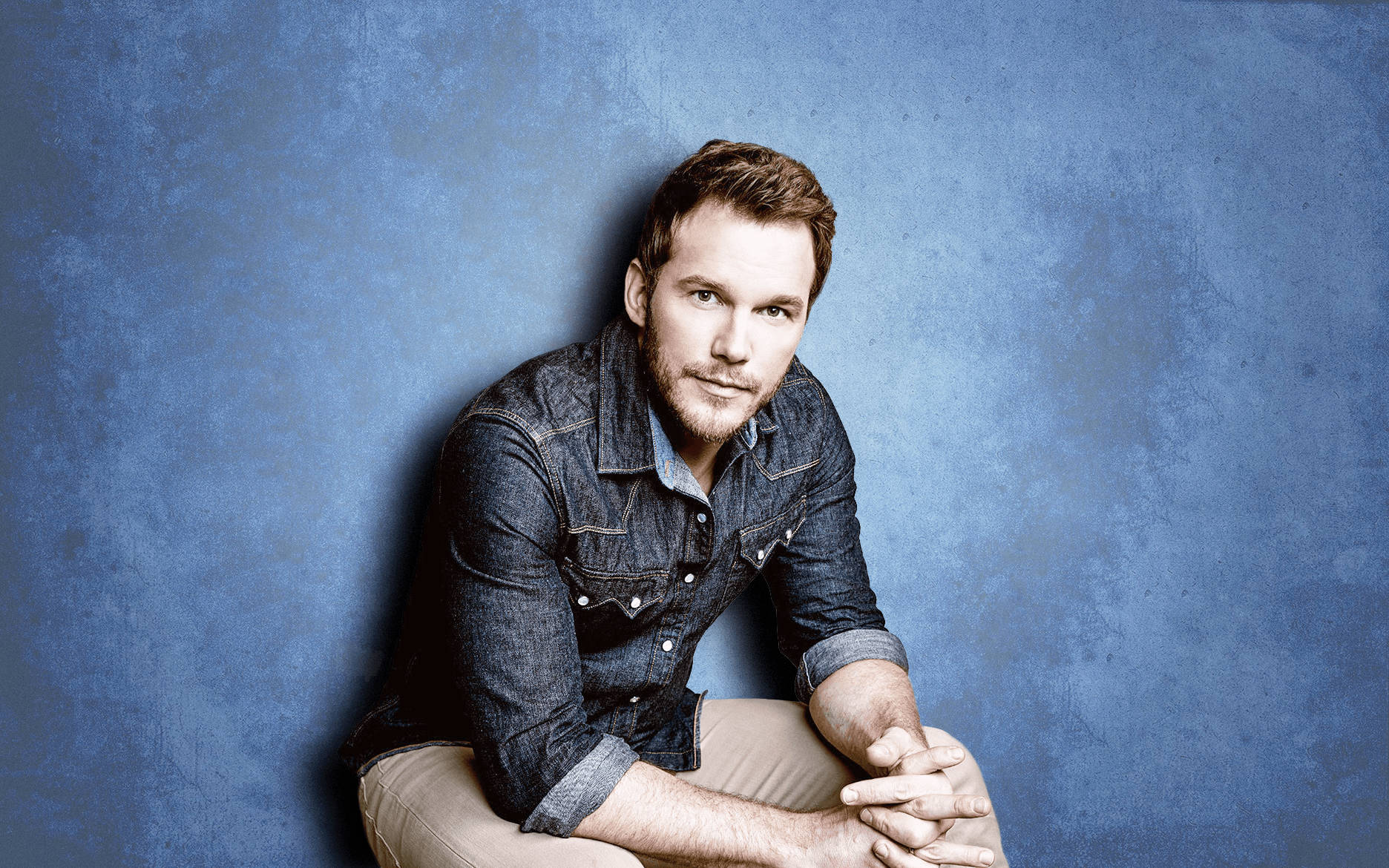 Chris Pratt 桌布 壁紙 — 一名身穿牛仔襯衫的男子靠著有紋理的藍色牆壁坐下，光線柔和，眼神直視。