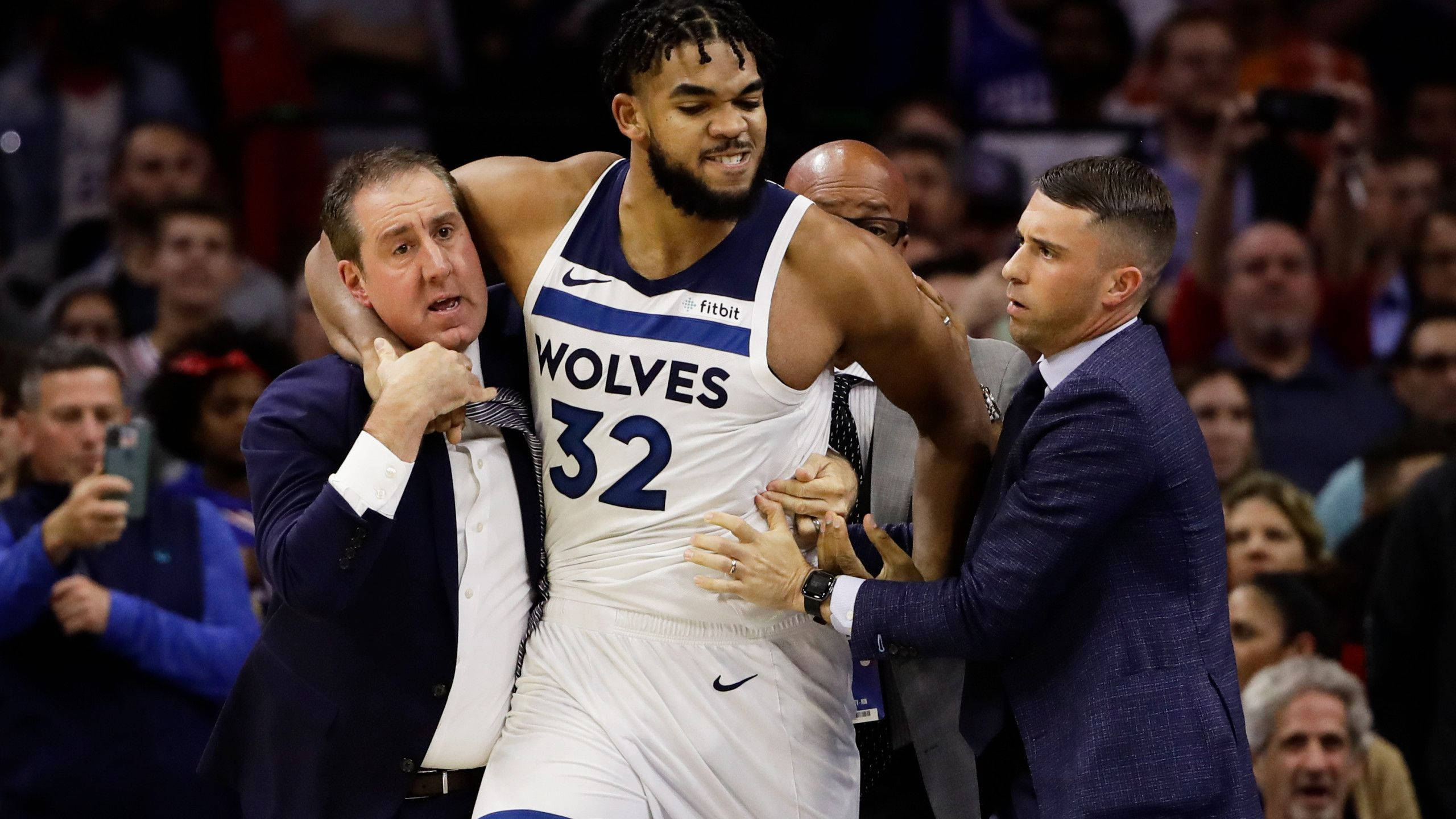 karl-anthony towns 桌布 壁紙 — 一名身穿狼隊球衣的籃球運動員在比賽期間被兩名穿西裝的男子扶離球場。