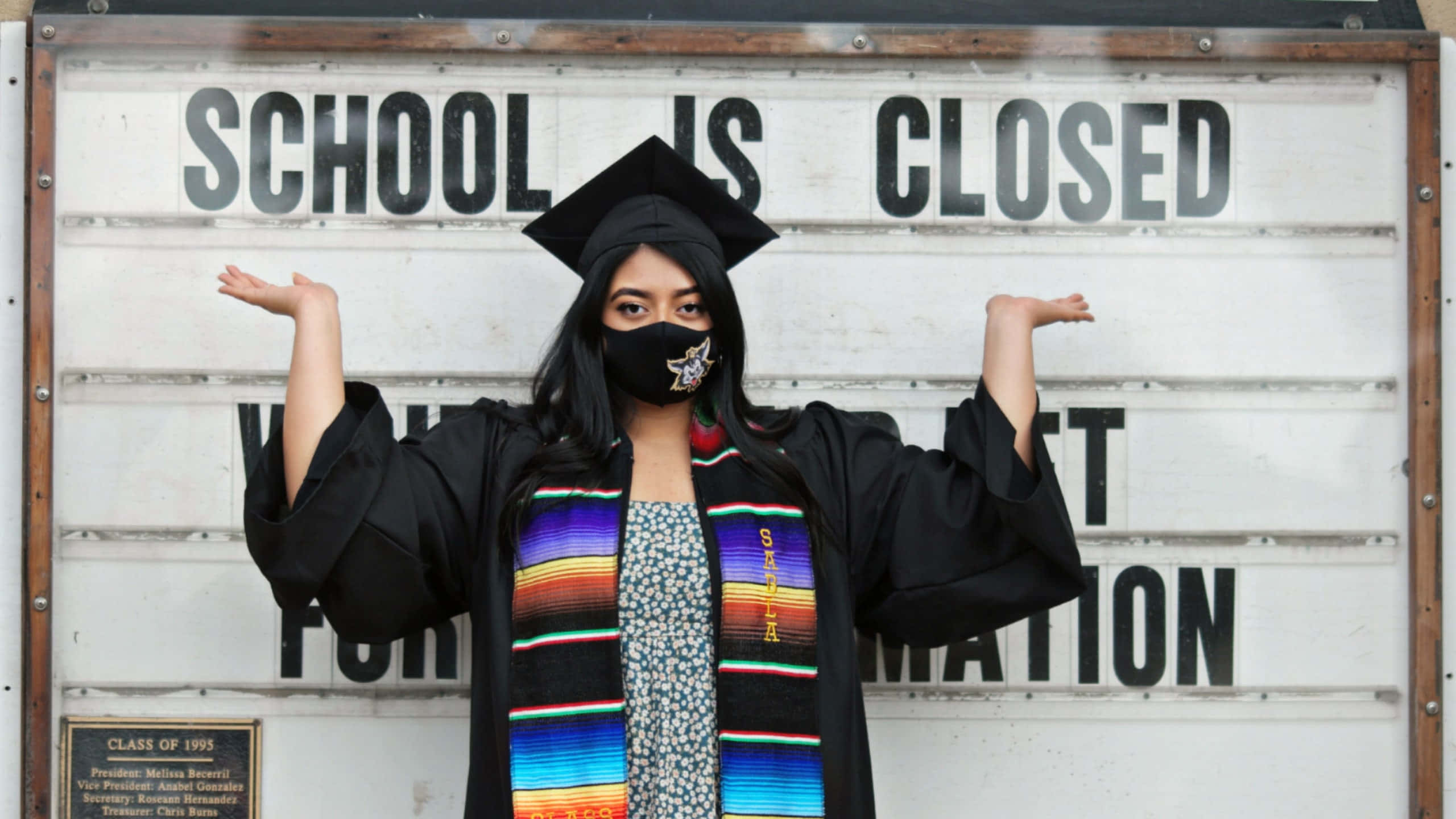 Graduation Ceremony 桌布 壁紙 — 一名身穿畢業袍、戴學士帽和彩色肩帶的畢業生，在「學校關閉」標誌前張開雙臂站立。