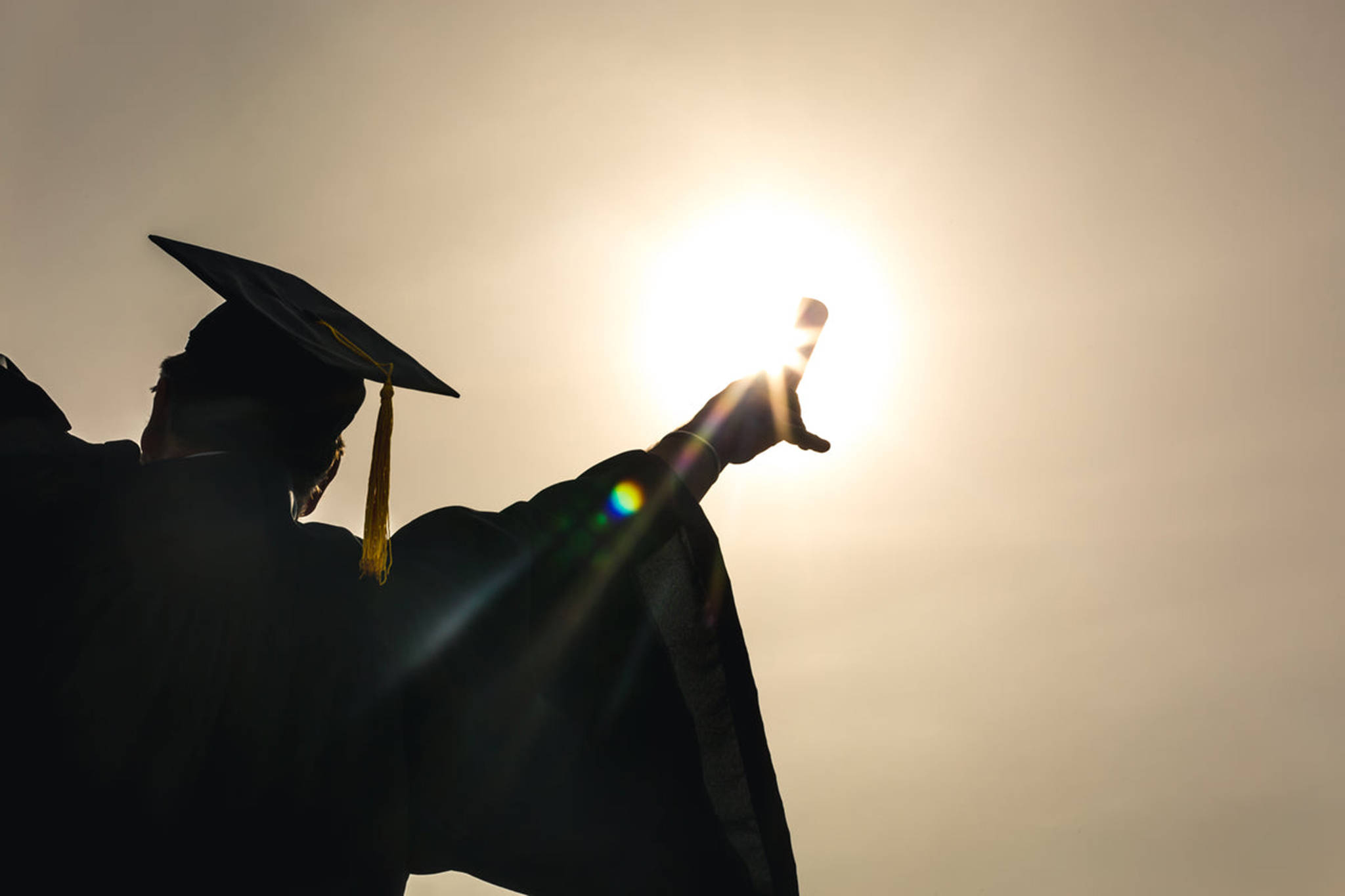 Graduation 桌布 壁紙 — 一名身穿畢業袍和學士帽的畢業生，在朦朧的天空下伸出手臂朝向太陽的剪影。