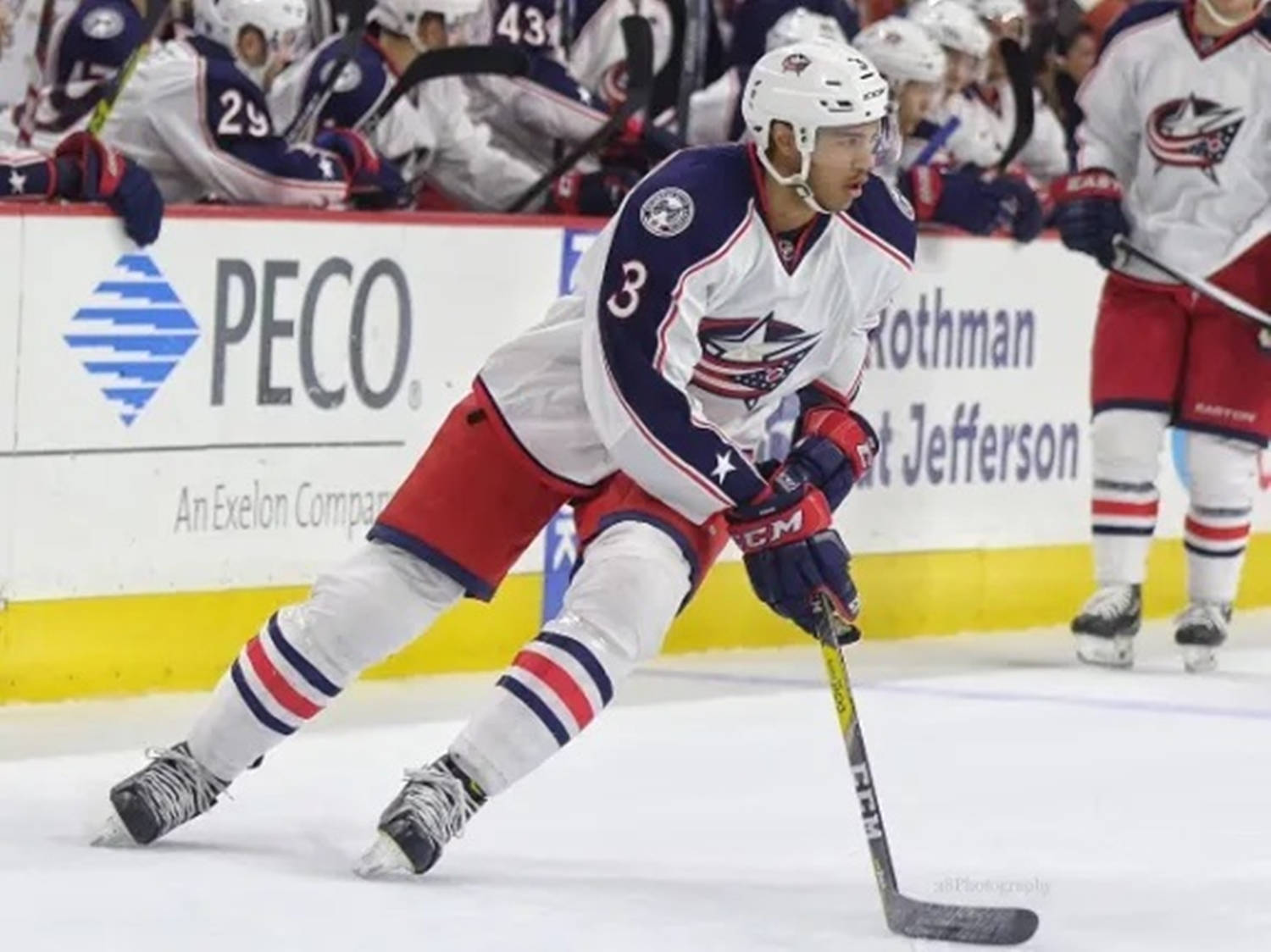 hockey 桌布 壁紙 — 一名身穿白、紅、藍色球衣的冰球運動員在比賽中與隊友和背景廣告一起，在冰上激烈地滑行並控球。