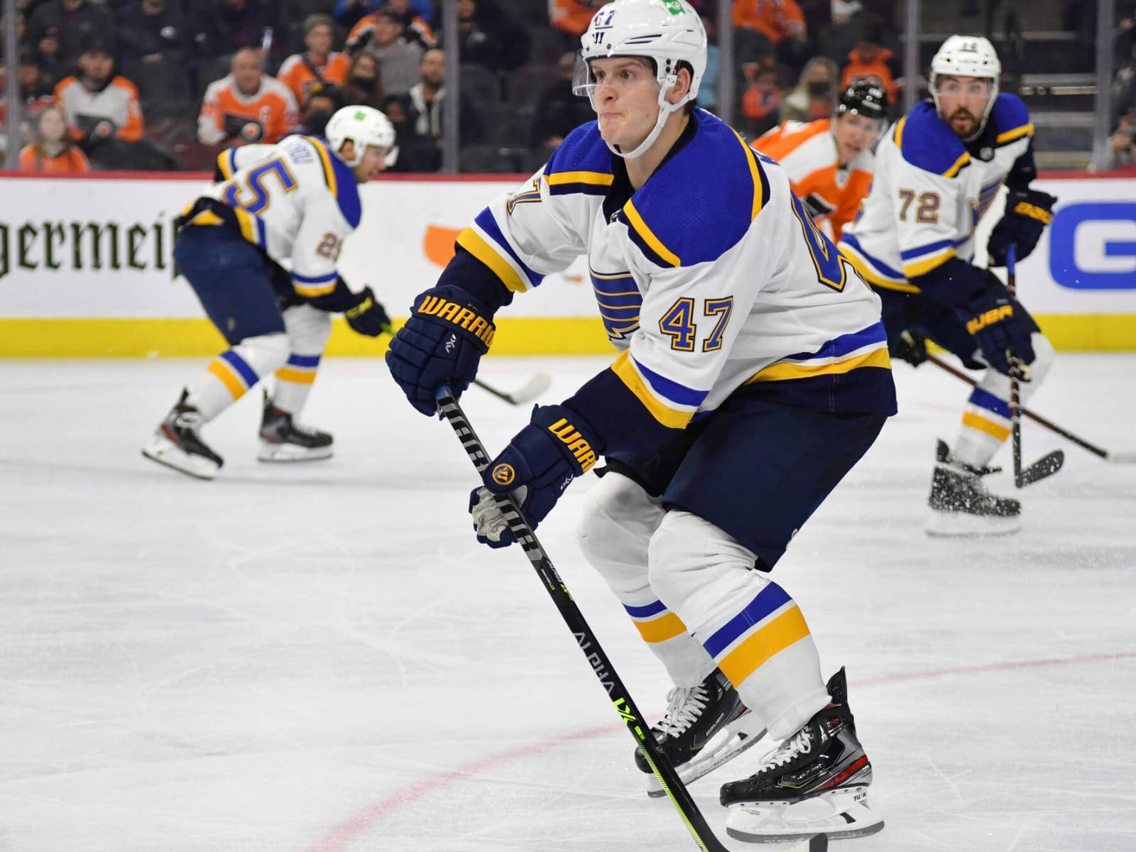 St Louis Blues 桌布 壁紙 — 一名身穿白、藍、金色球衣的冰上曲棍球運動員在比賽中與隊友和對手激烈地在冰上滑行。